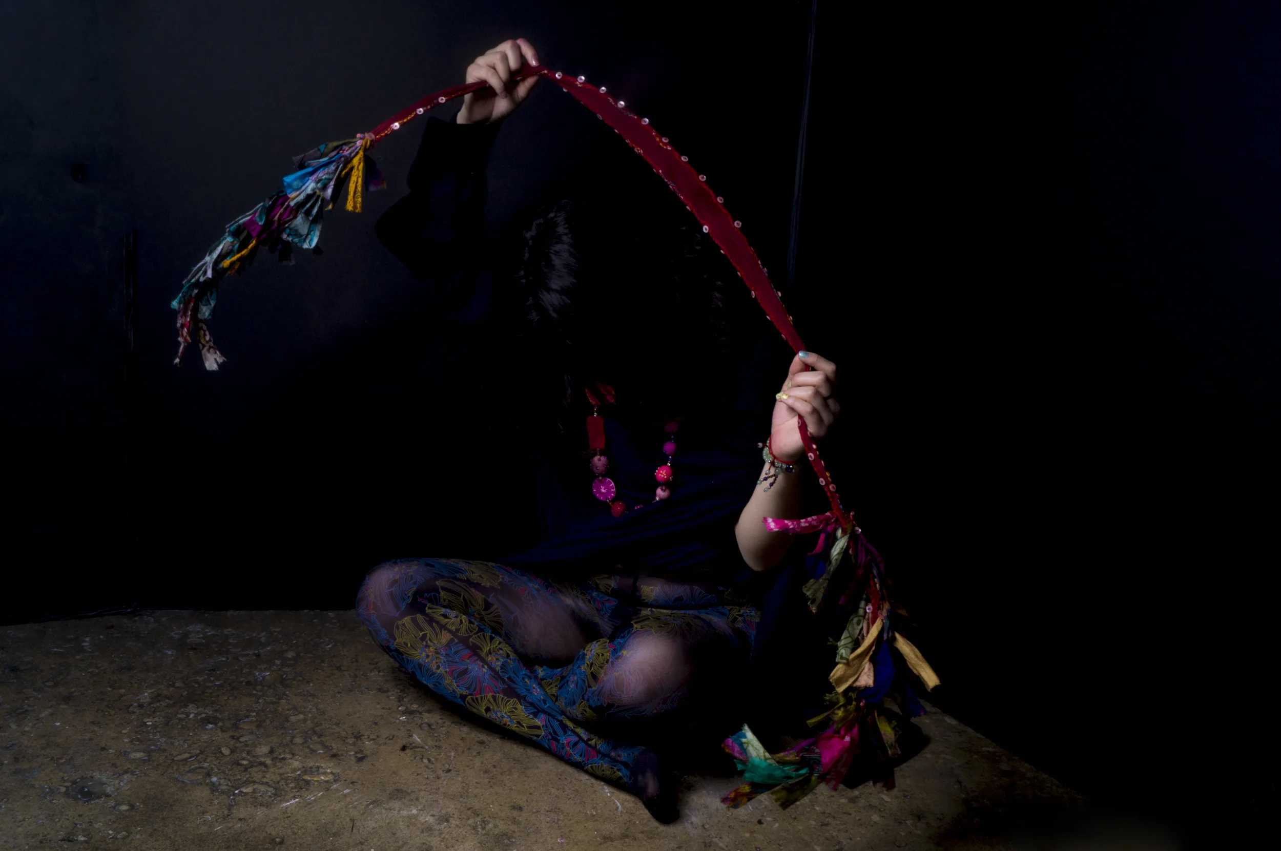 A person kneeling on the ground, holding a colorful, decorated headband over their face, dressed in vibrant patterned clothing against a dark background.