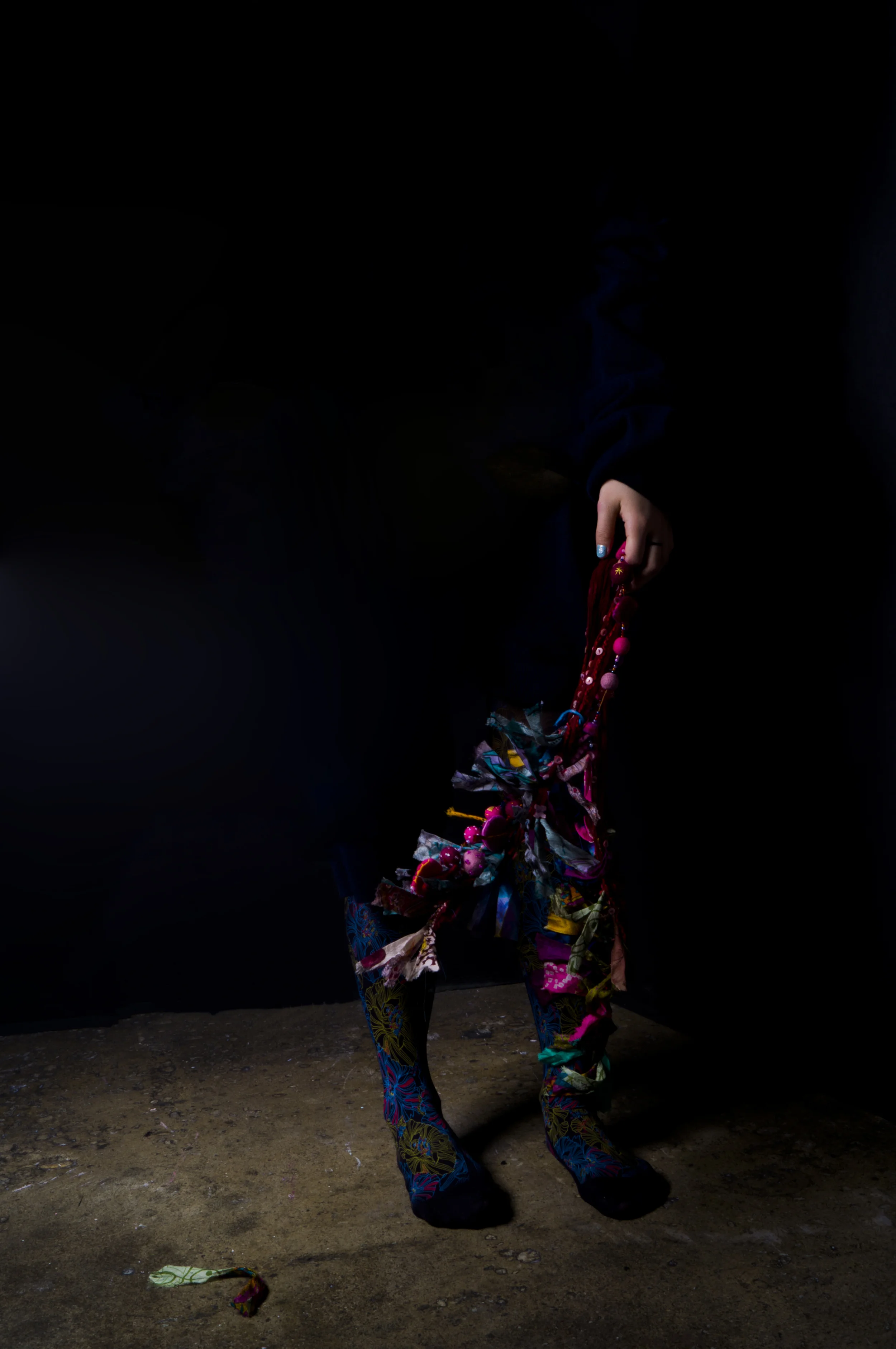 Person holding colorful fabric and jewelry, standing in a dark space with bright patterned clothing, illuminated to highlight their attire.