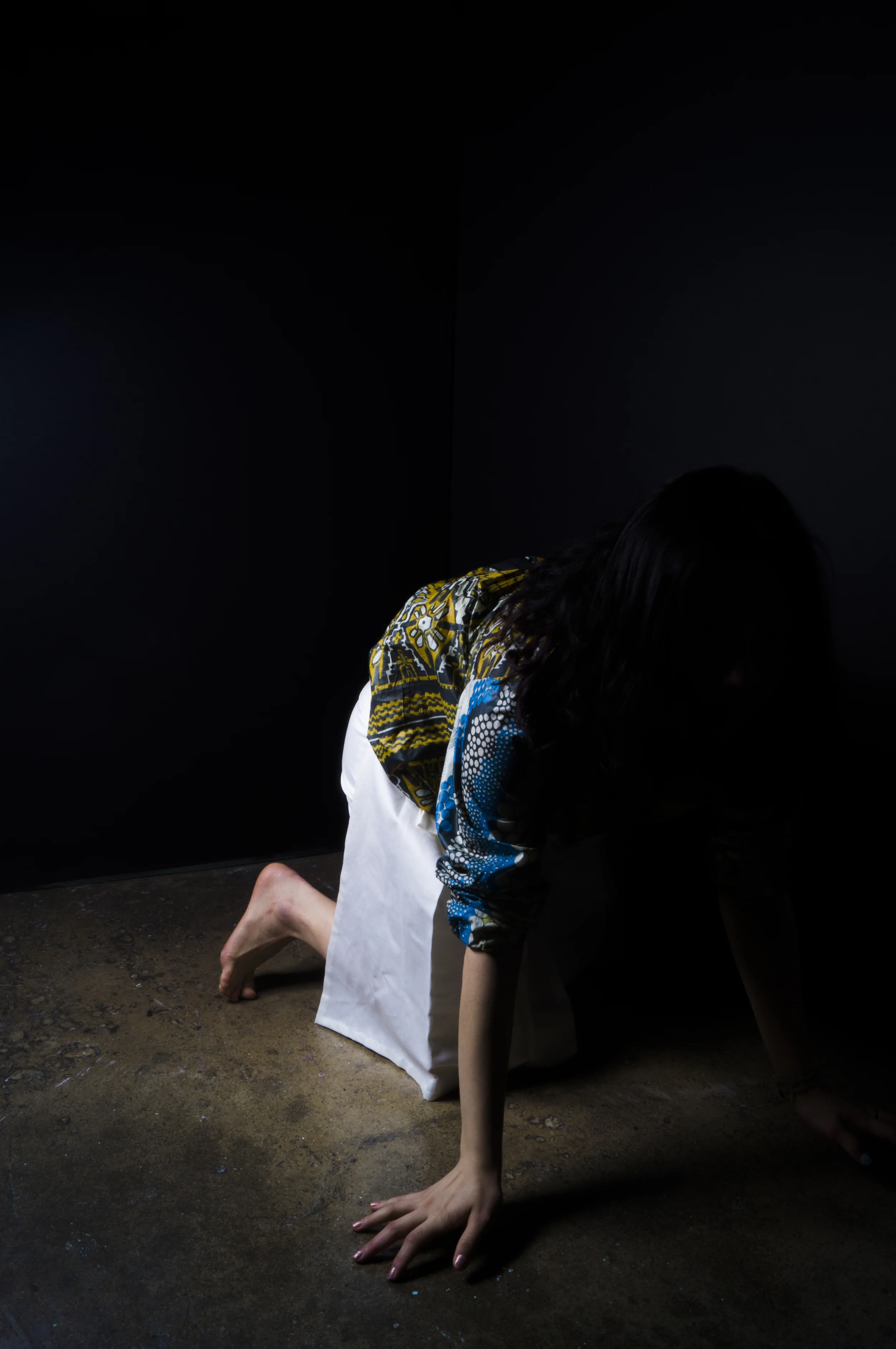 Person in colorful clothing and a white apron on hands and knees on a dirt floor, against a black background.