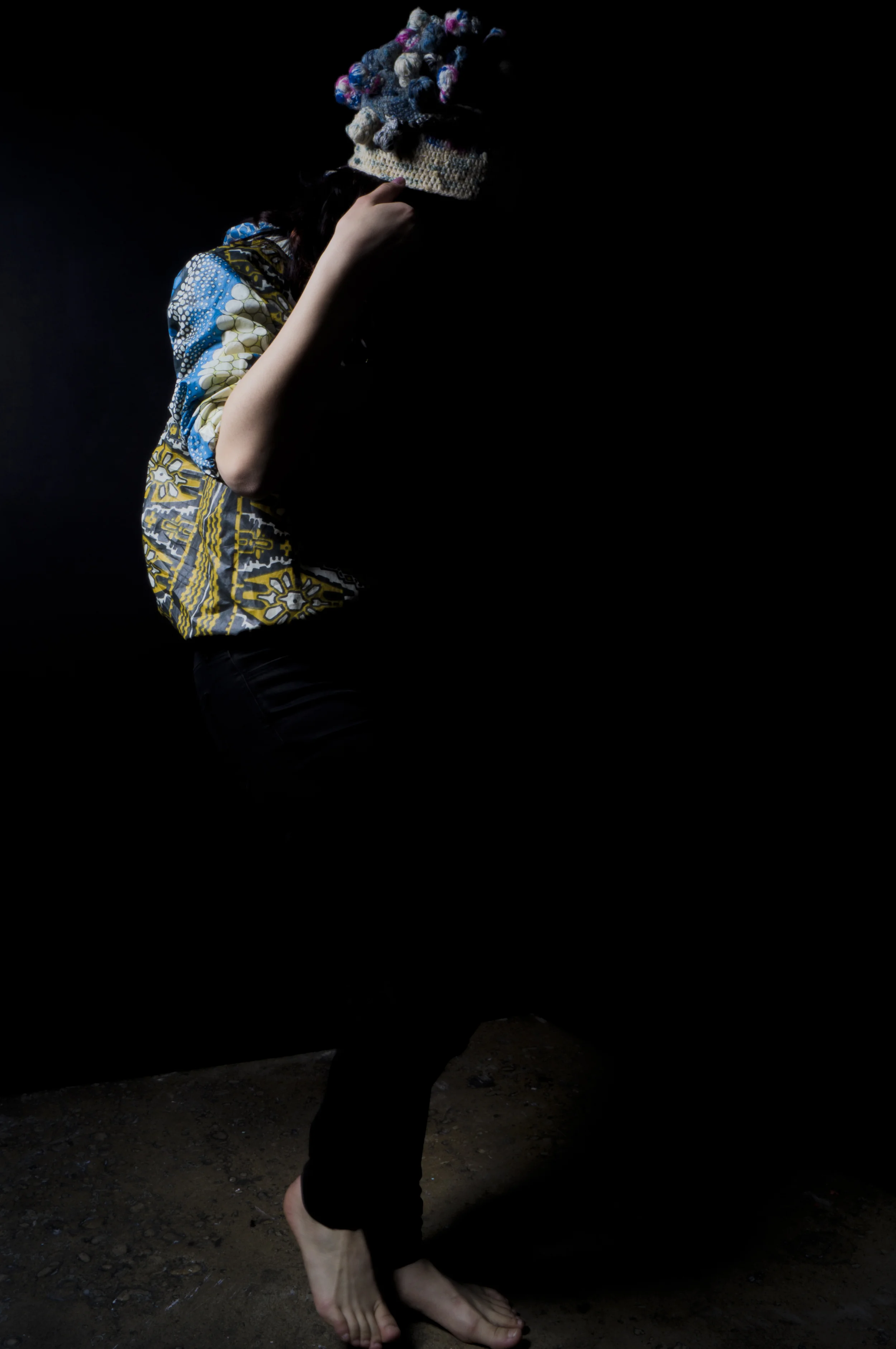 A person walking outdoors on dirt ground, wearing a colorful patterned shirt and a multicolored knitted hat with pom-poms, seen from the side with no visible face.