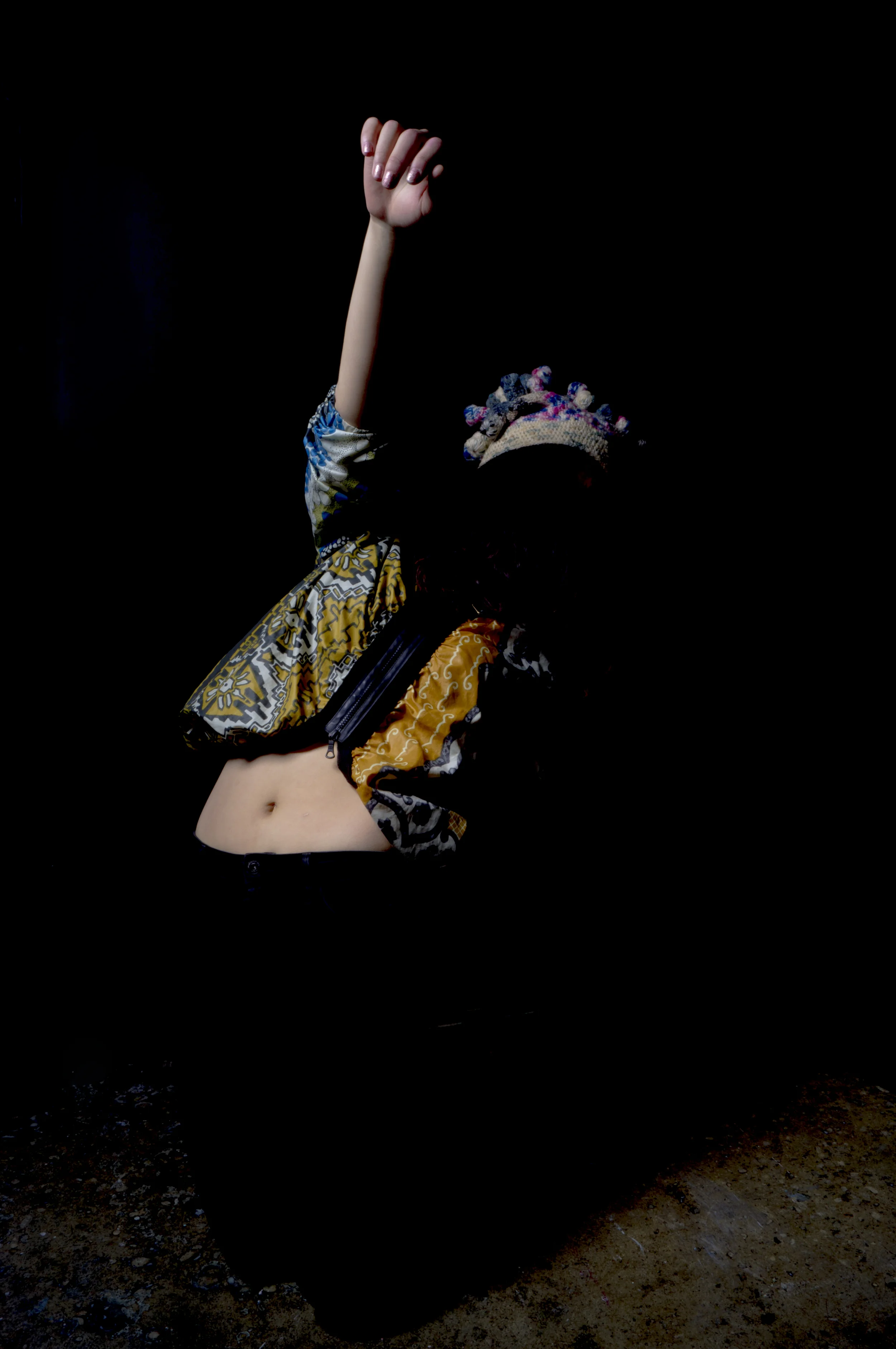 A person with a patterned jacket and a colorful knitted hat a their face is outside, with their right arm raised and their hand clenched into a fist.