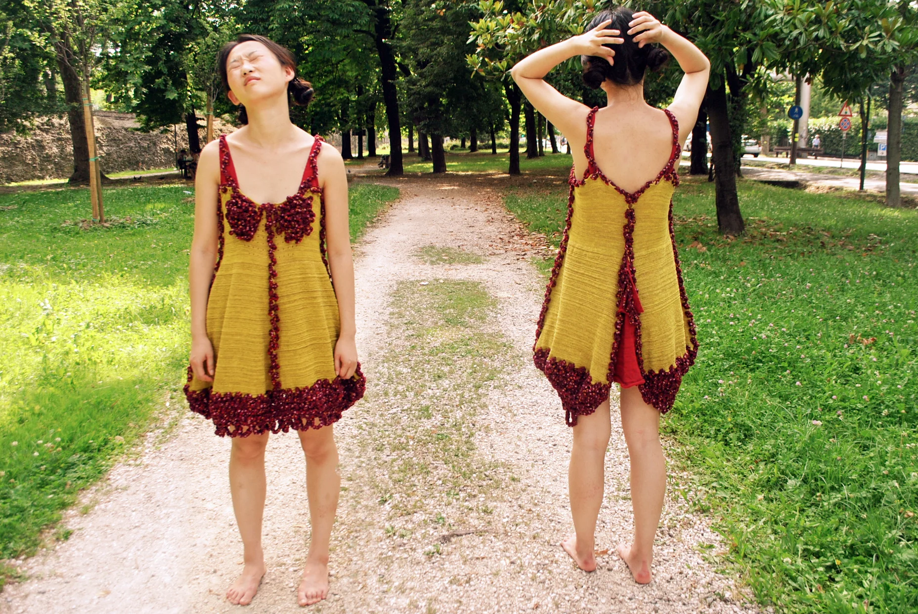 Two women in yellow dresses with red trim and embellishments, standing barefoot on a gravel path in a park with green trees and grass, one facing forward with eyes closed and the other turned away touching her hair.