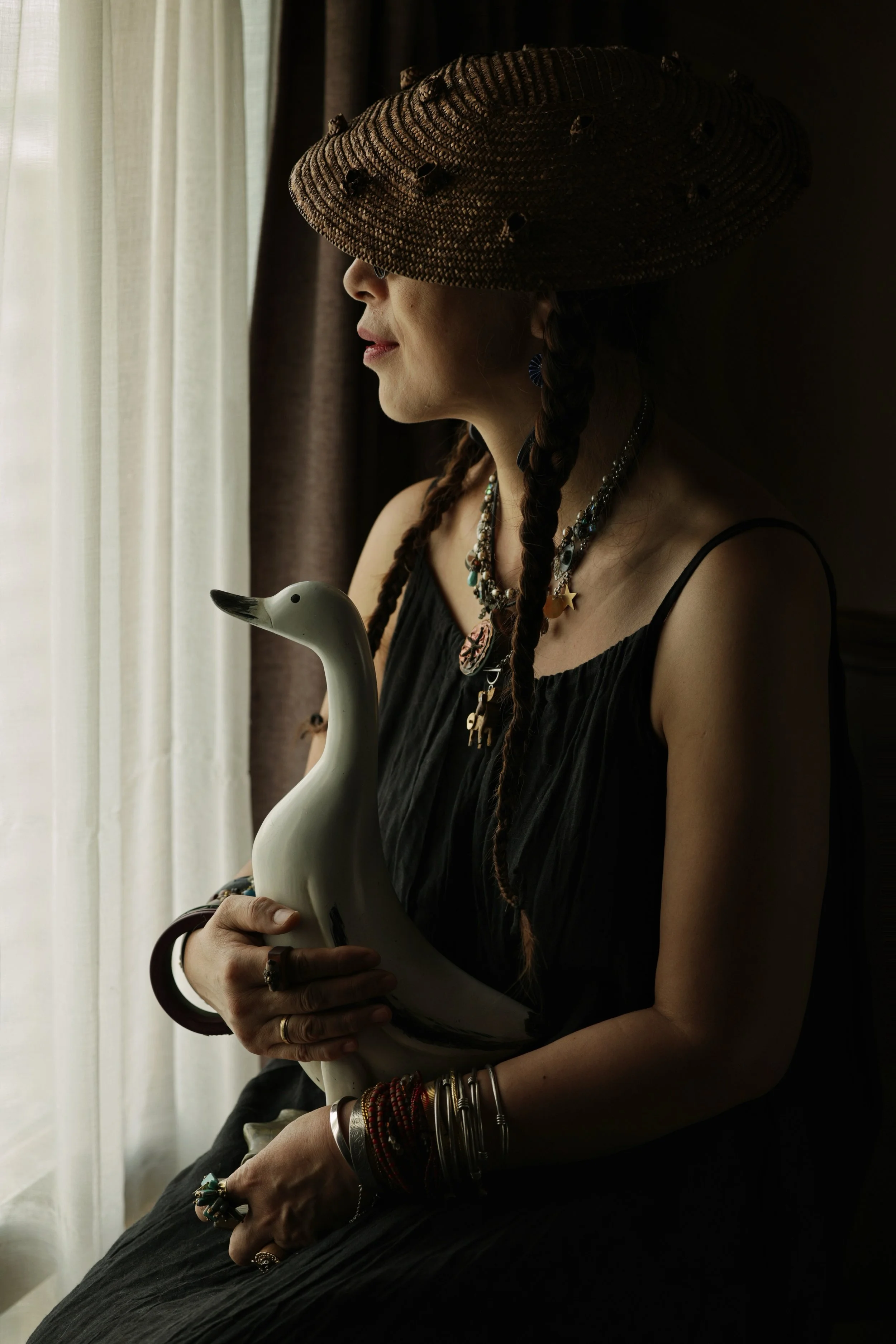 A woman with braided hair, wearing multiple necklaces and bracelets, is holding a ceramic swan figurine. She is standing by a window with sheer curtains, and is wearing a large, woven hat that obscures her face.