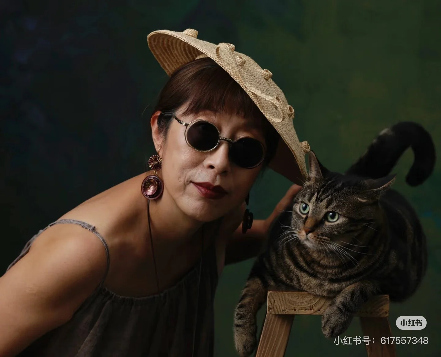A woman wearing a straw hat with decorative elements, sunglasses, and earrings poses with a tabby cat that is sitting on a small wooden stool, against a dark green background.