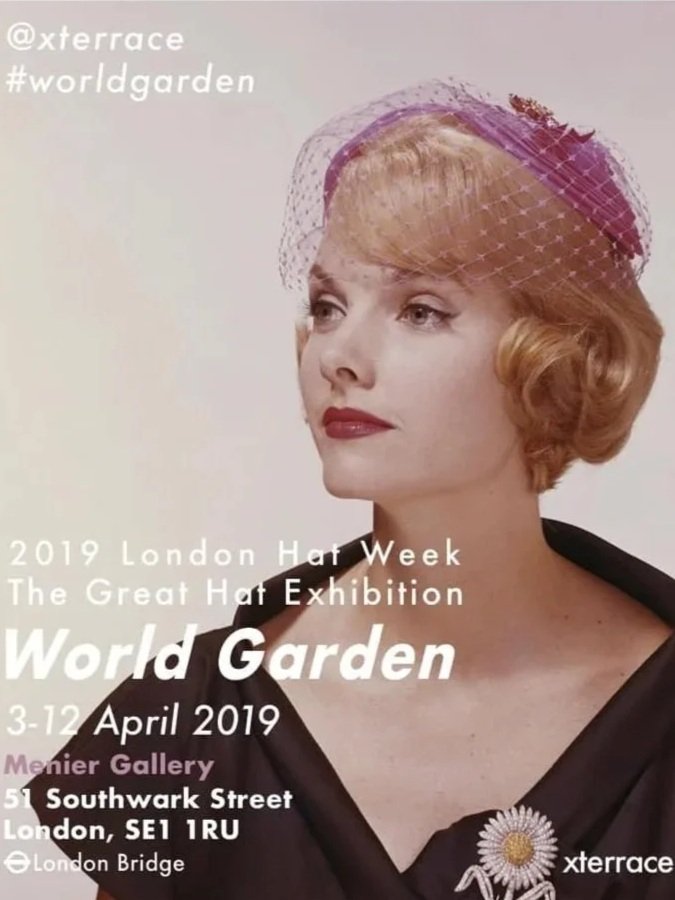 A woman with short, wavy red hair wearing a pink hat with a veil, black shawl or coat, and a daisy pin. Promotional poster for the 'World Garden' hat exhibition in London, April 3-12, 2019.