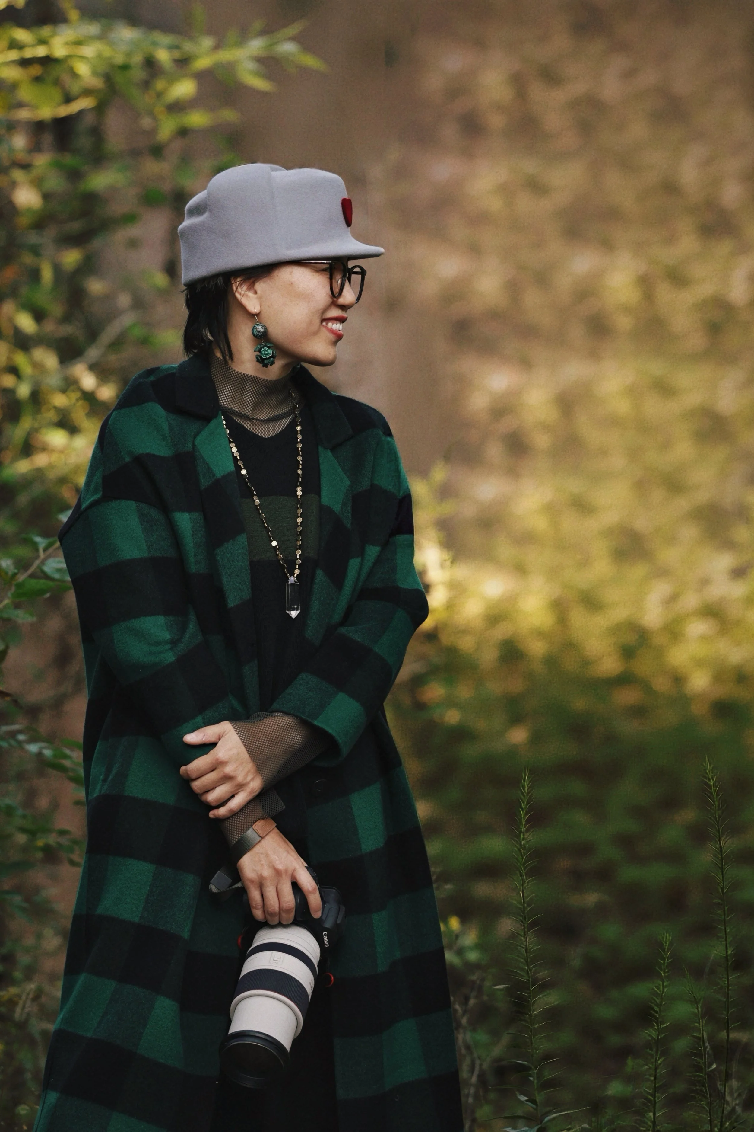 A woman wearing a grey hat, glasses, and green and black checkered coat, standing outdoors with blurred autumn foliage background, holding a Canon camera with a large telephoto lens.