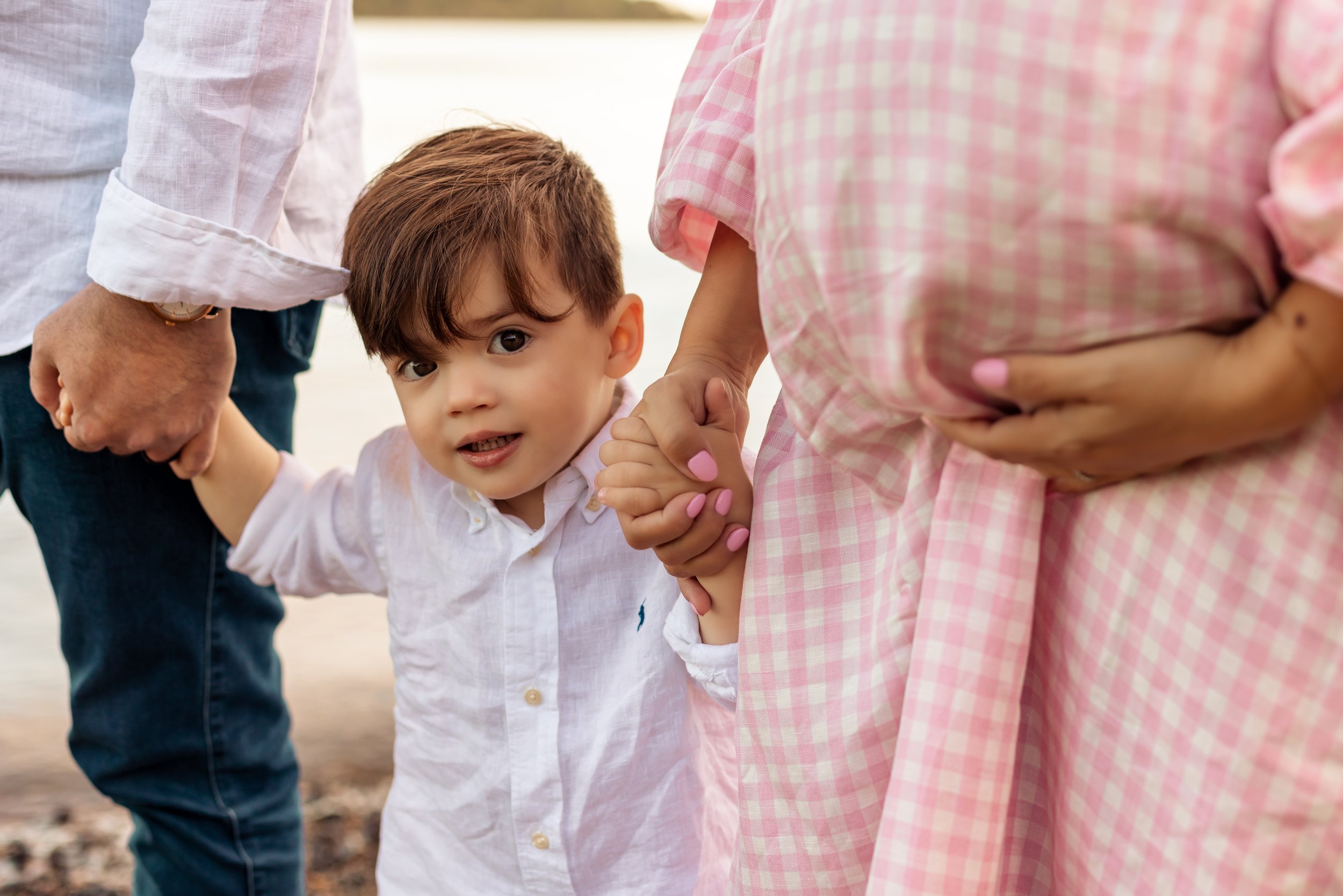 A young child holds hands with two adults, one wearing a white shirt and the other in a pink checkered dress, with a scenic outdoor backdrop.