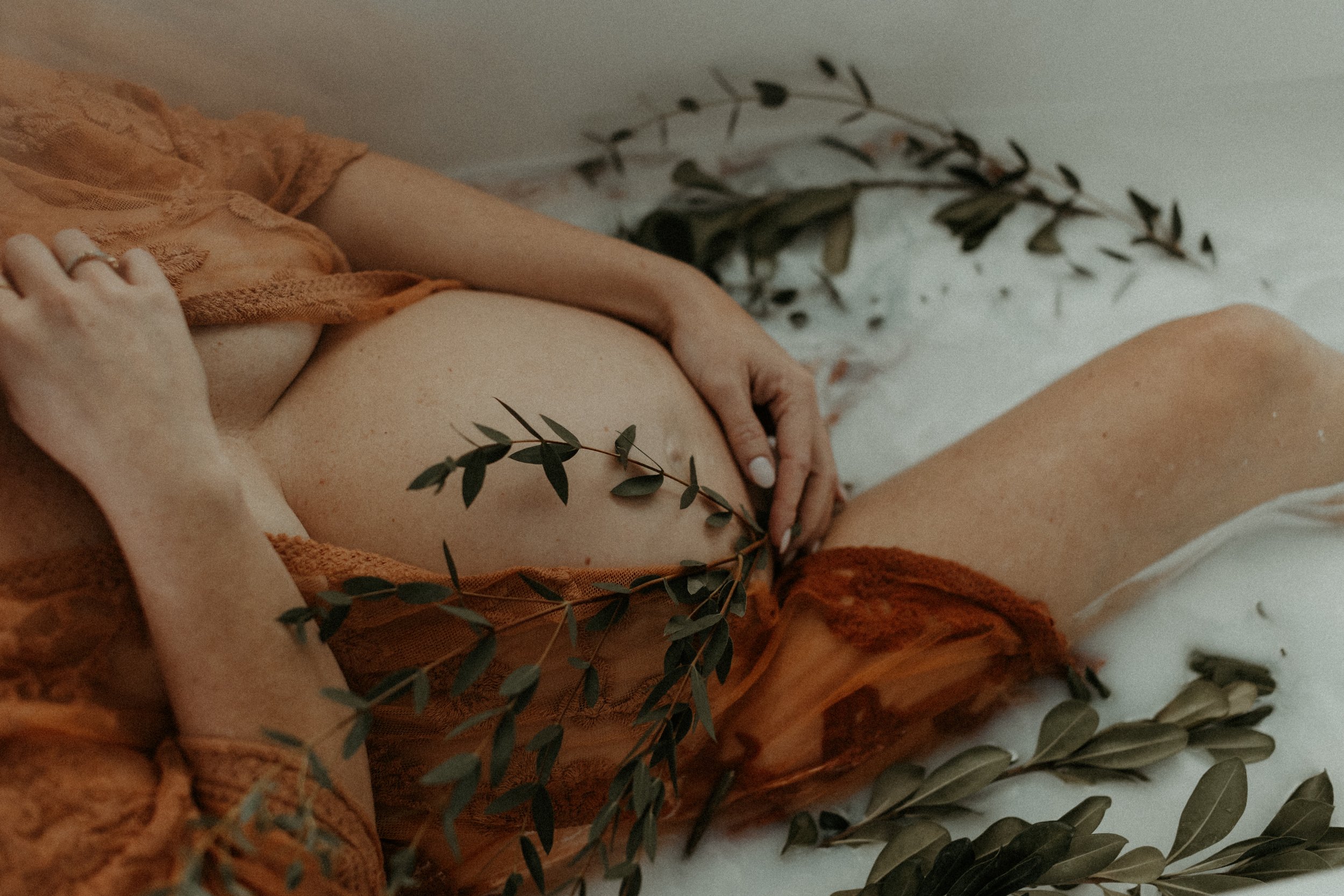 Pregnant woman in lace dress with greenery, relaxing in milk bath.