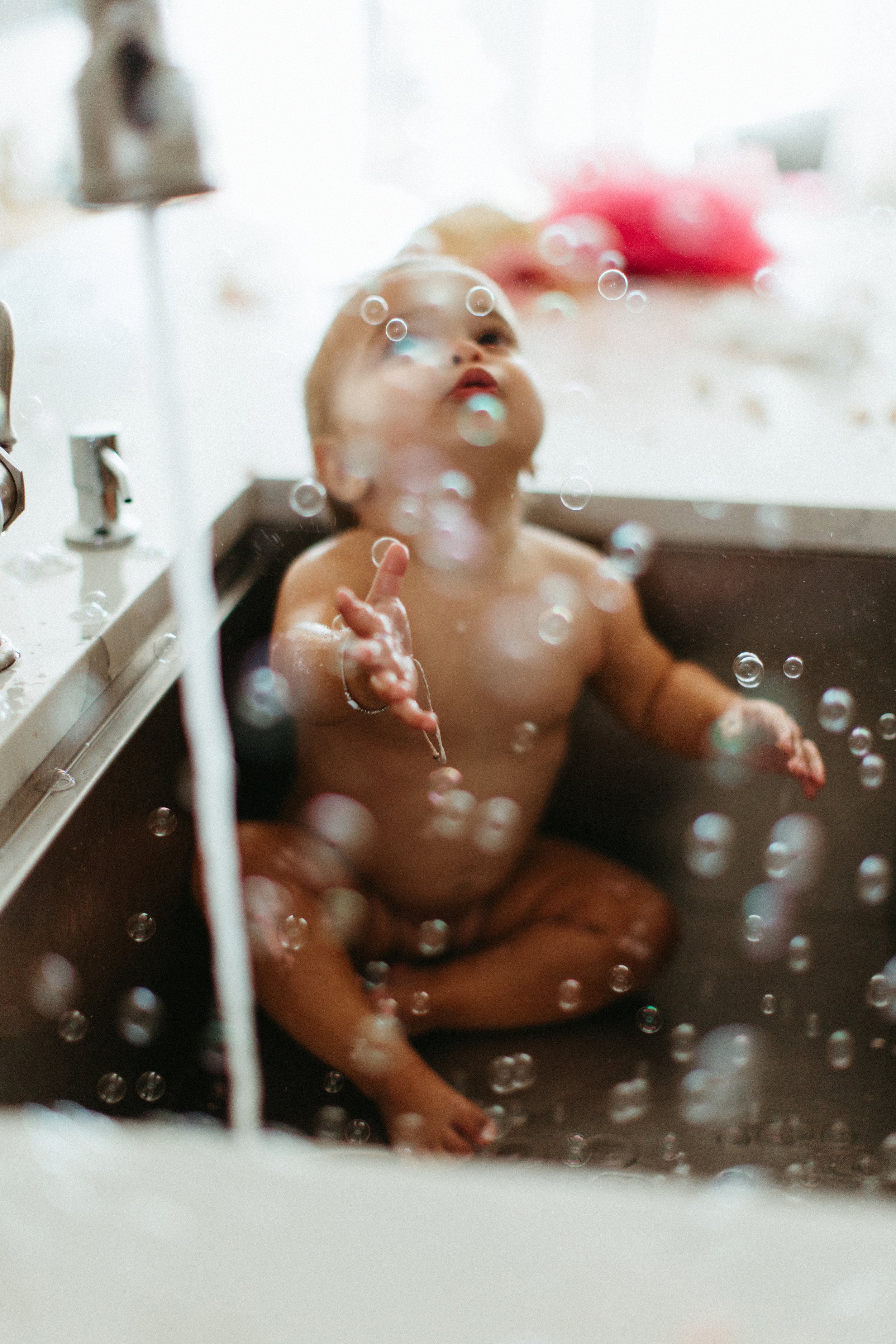 Baby playing with bubbles in a kitchen sink under running water.