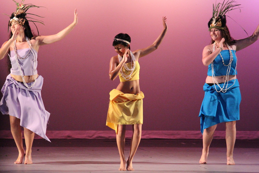  Three girls dancing a pacific islander dance. They are each wearing lilac, yellow, and blue. 
