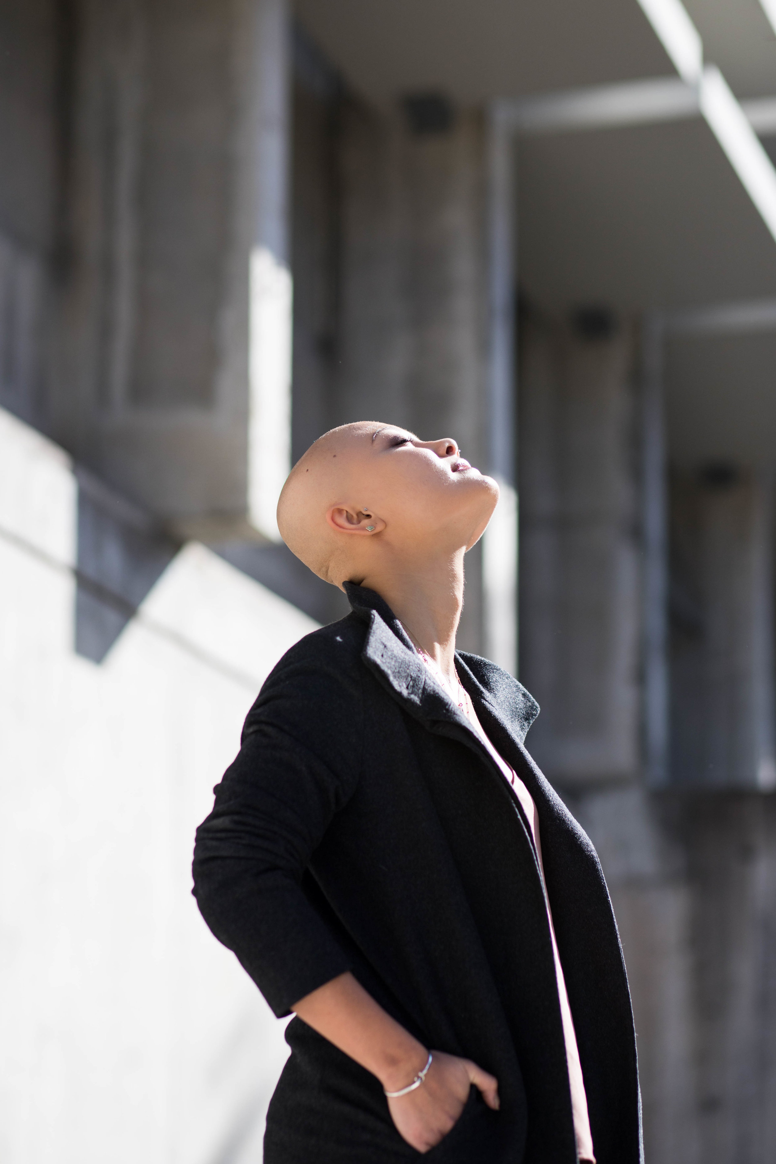  Woman wearing black coat with head tilted back and face up to sun against concrete budding 