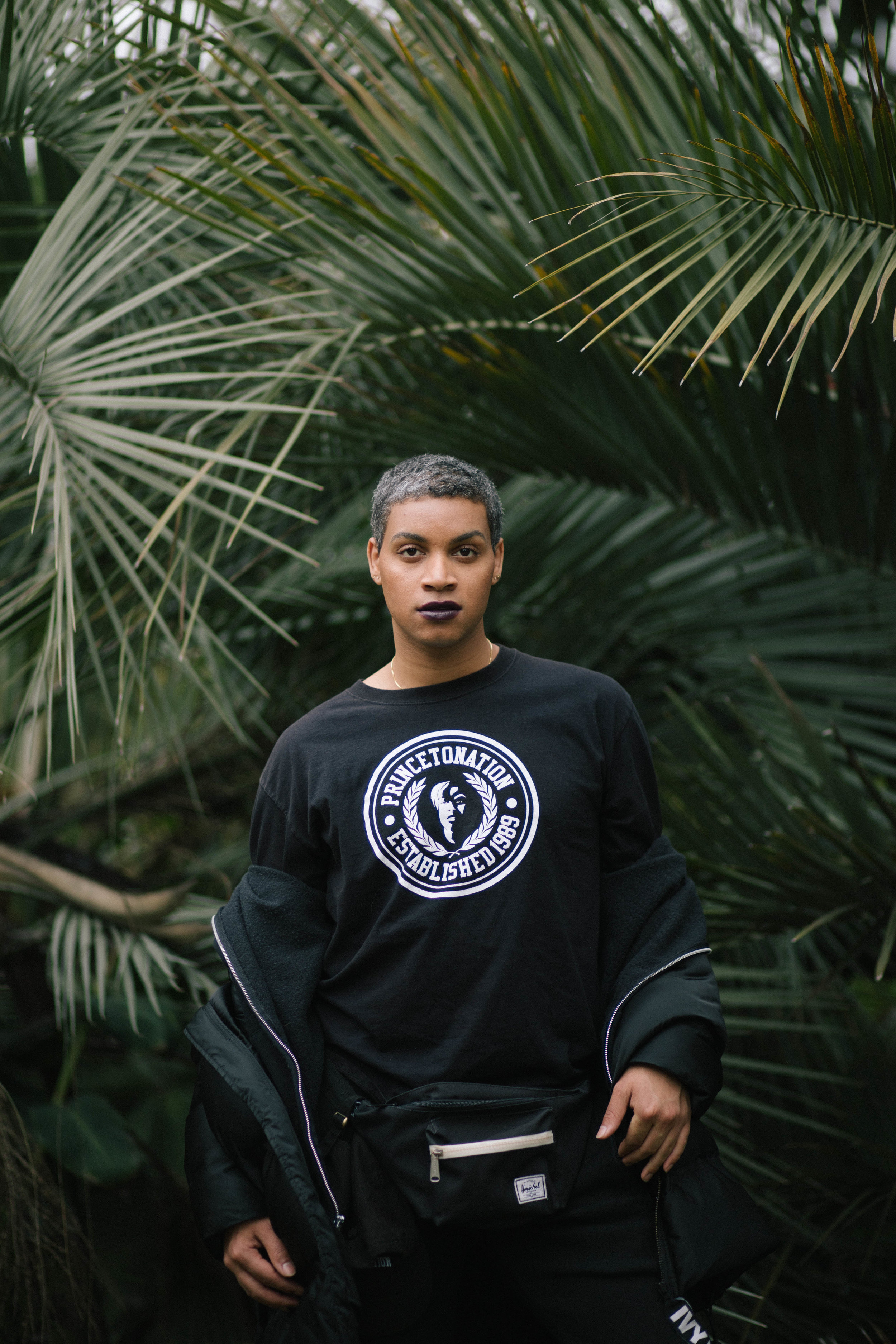  Man surrounded by palm fronds wearing back Princetonation crewneck and black puffer coat 