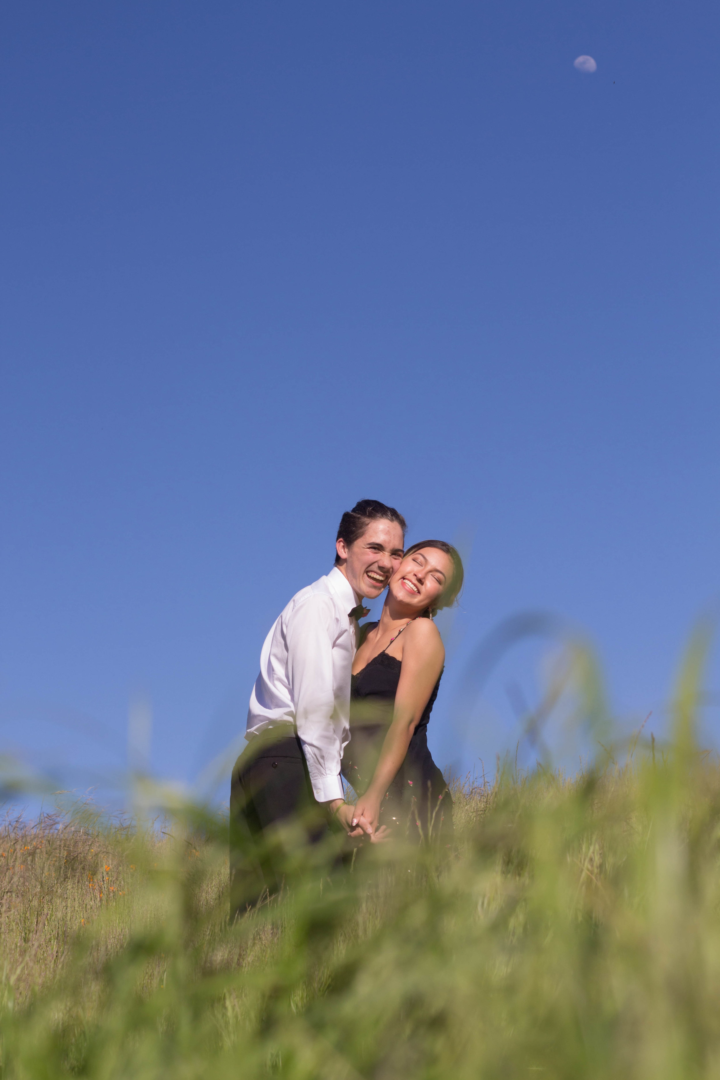  Couple holding hands and looking at camera leaning to the left. Grass in the foreground. Blue sky in the background with the moon faintly visible. 