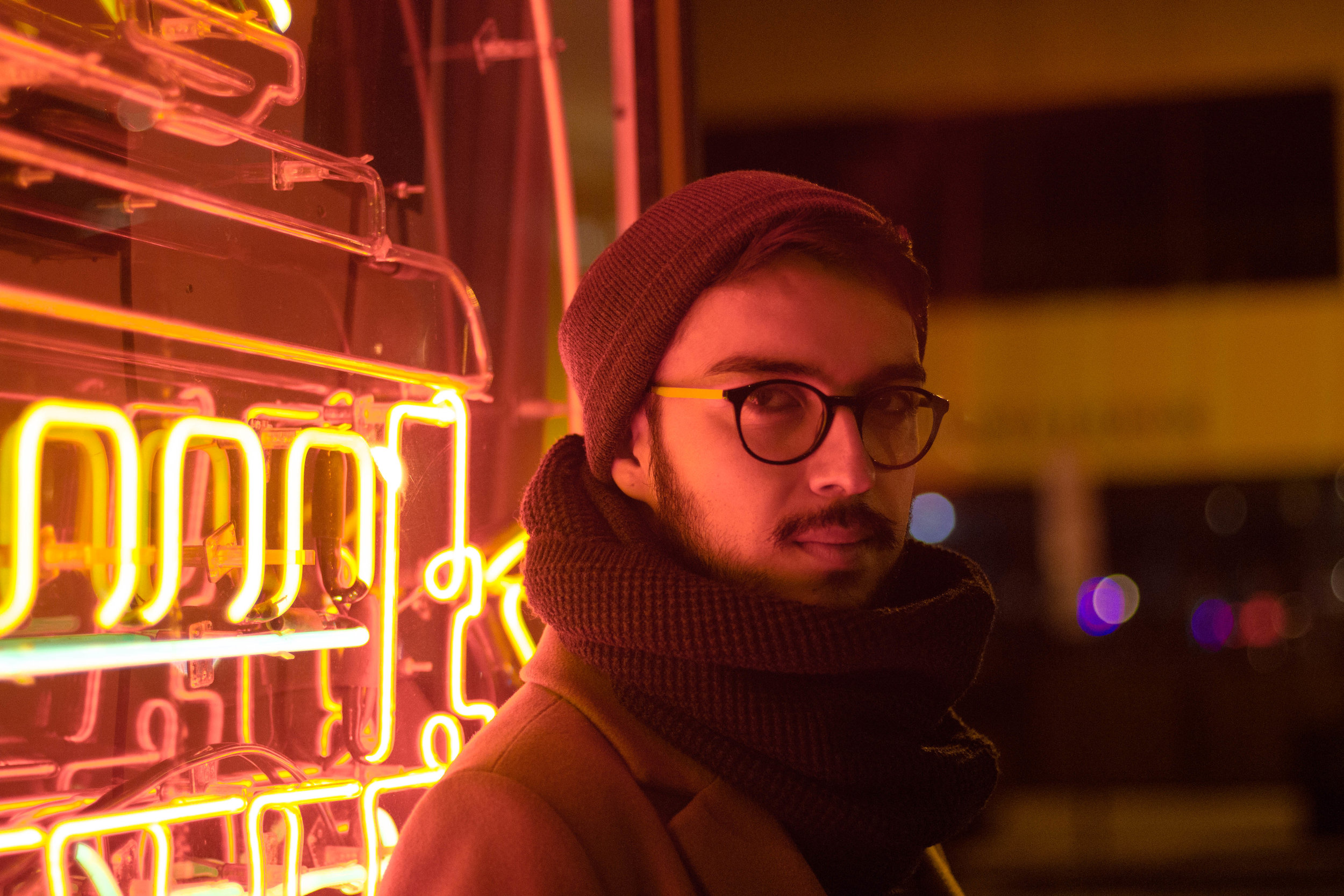  Man standing in front of red and orange neon sign. He is wearing glasses, a beanie, and a thick scarf. 