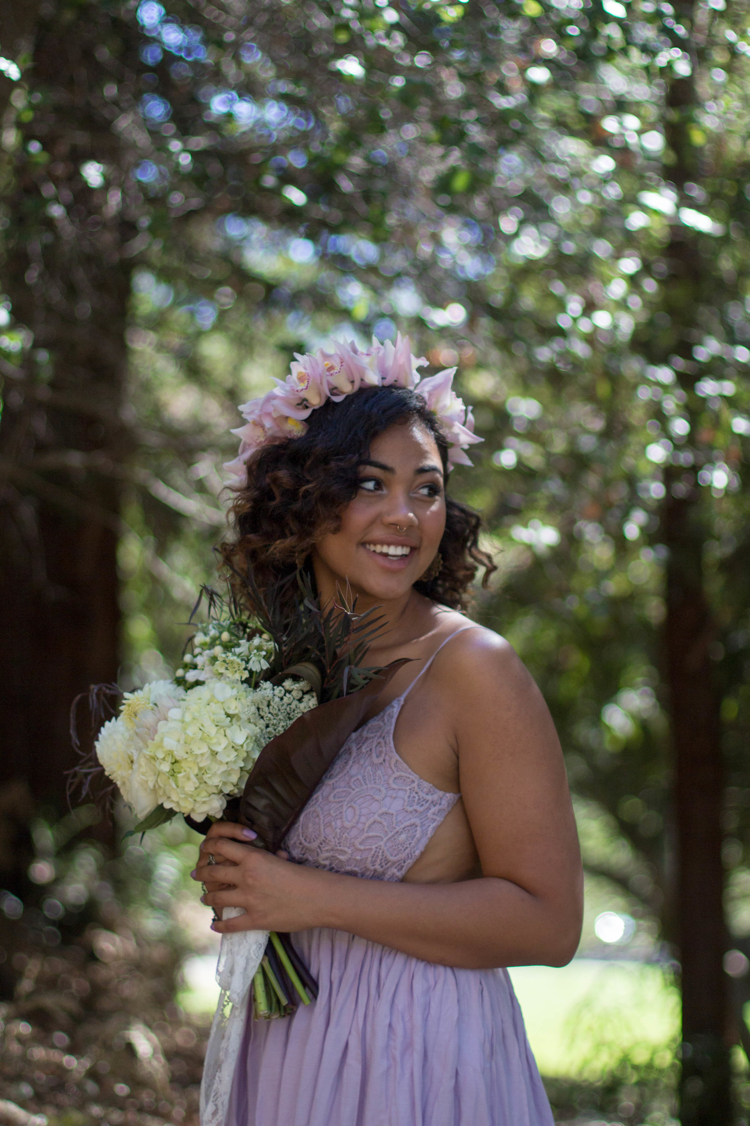  Woman wearing lilac dress and crown of pink and white flowers holding a bouquet of white flowers in a forrest 