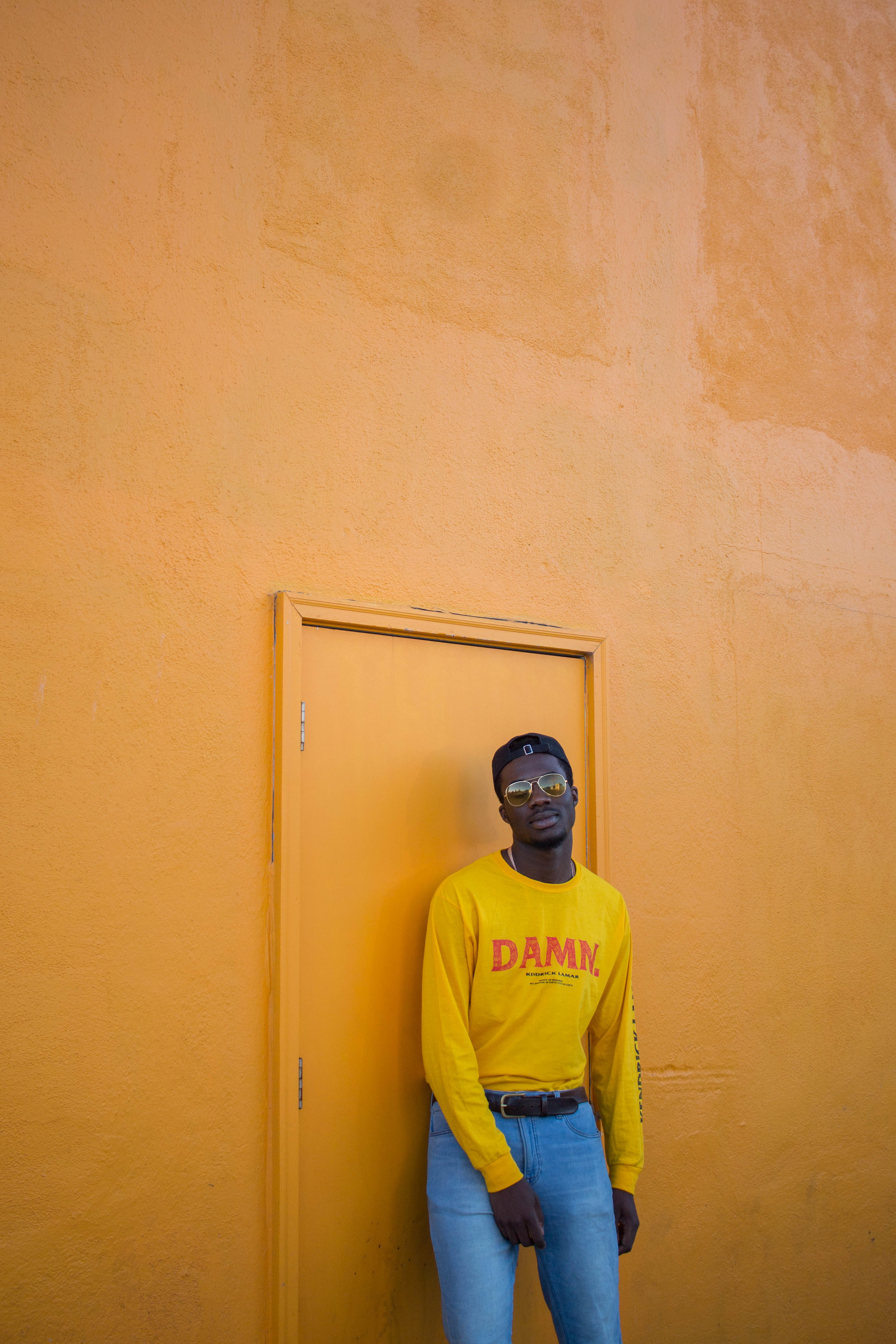  Man standing in front of yellow wall with a door wearing yellow shirt and light blue jeans. 