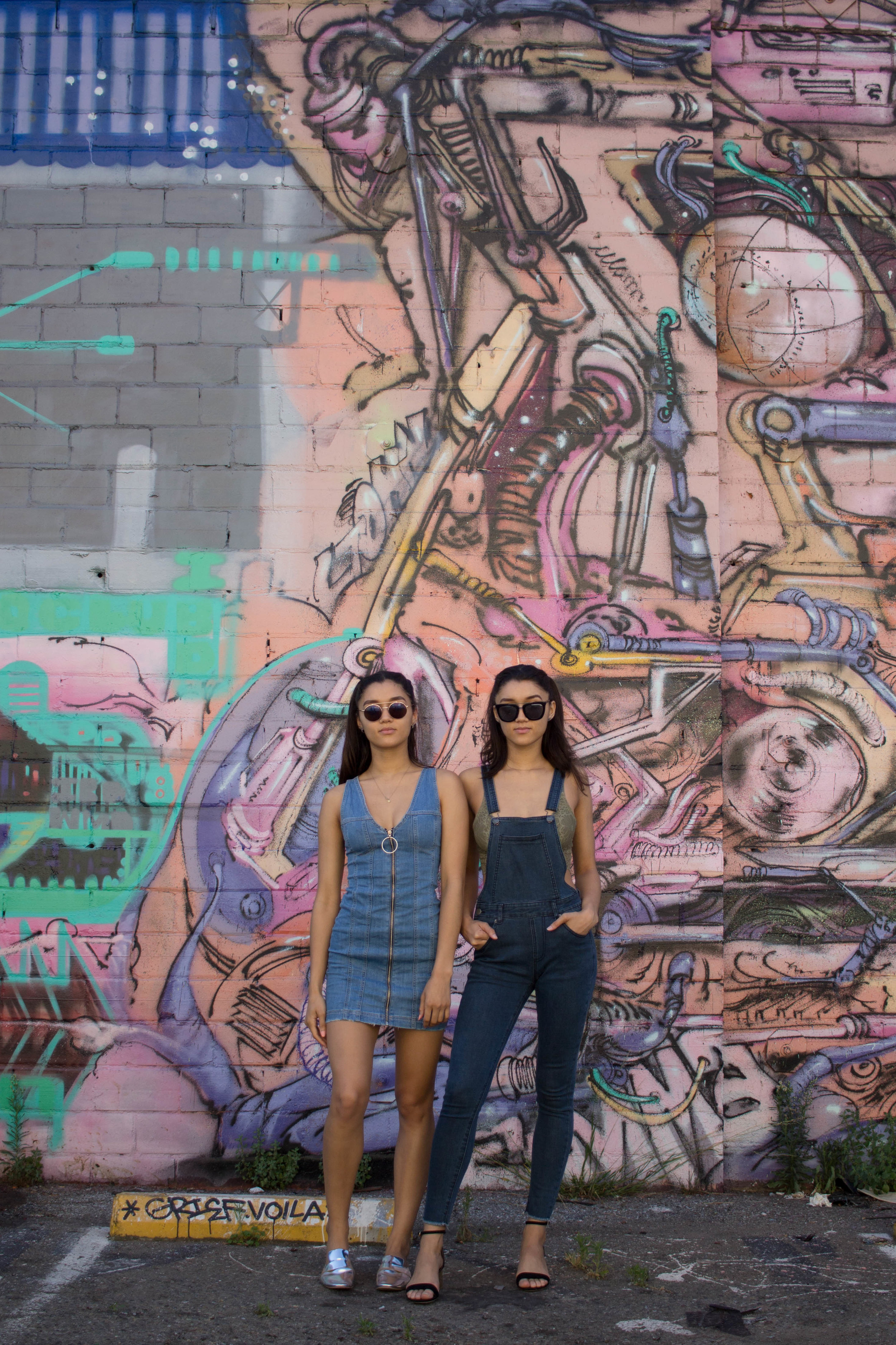  Twins standing infant of pink and pastel graffiti-ed wall. Woman wearing sunglasses and one wearing a denim dress and the other wearing dark denim overalls. 