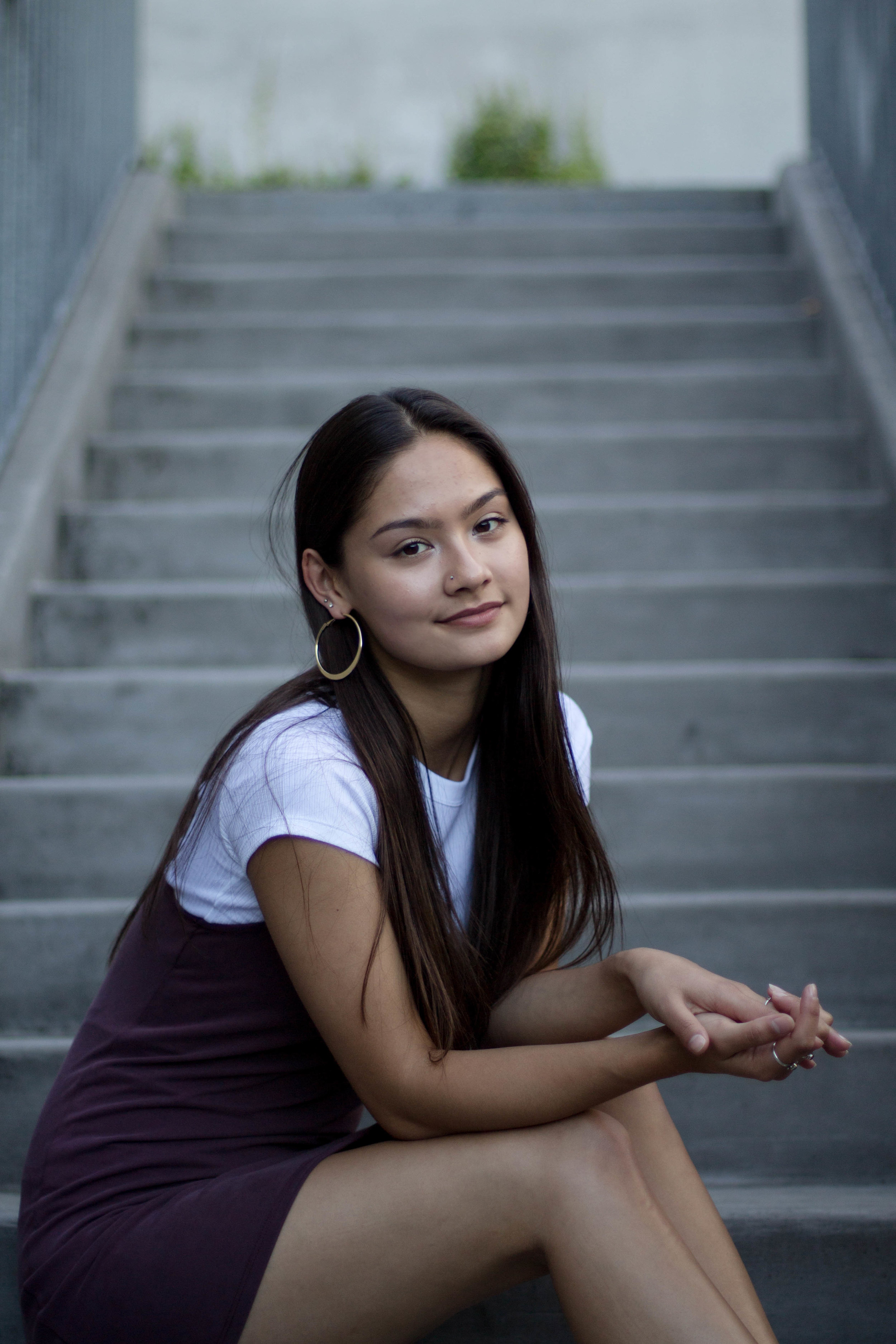  Girl sitting on concrete steps wearing white t-shirt with dark overall dress on top. She has her elbows resting on her knees and is looking at the camera over her right shoulder. 