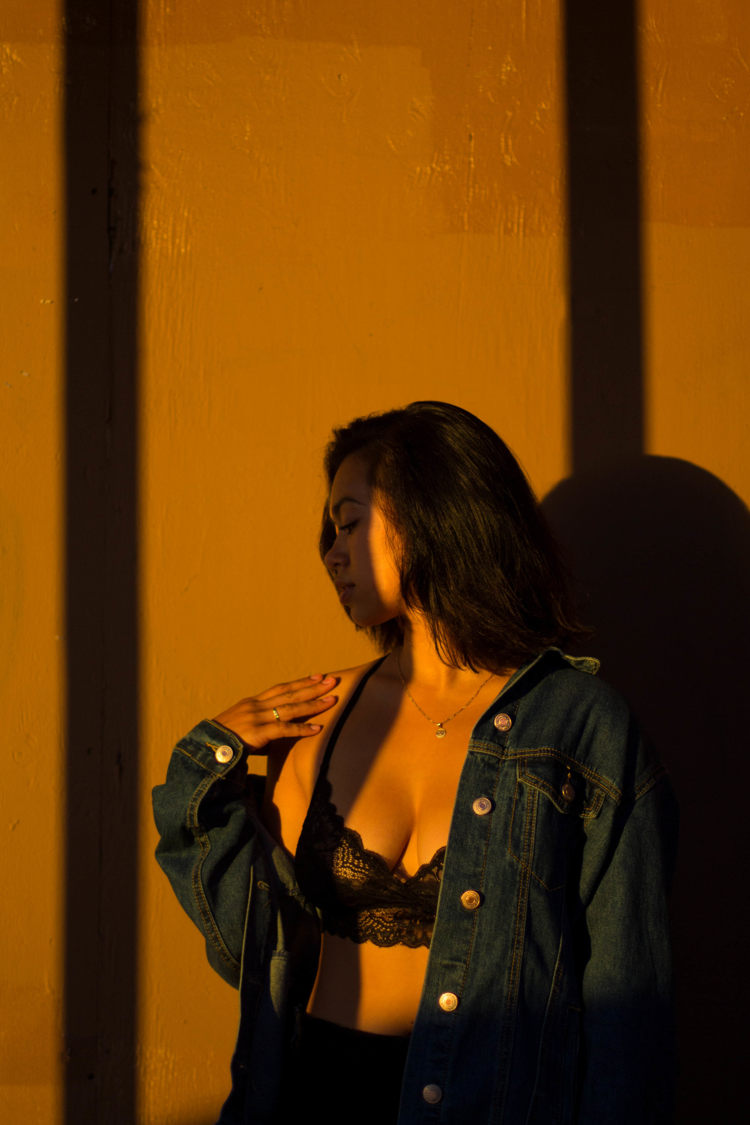  Woman standing in front of a yellow wall looking to her side with her hand on her shoulder and two linear shadows across the image. She is wearing a denim jacket and black bralette. 
