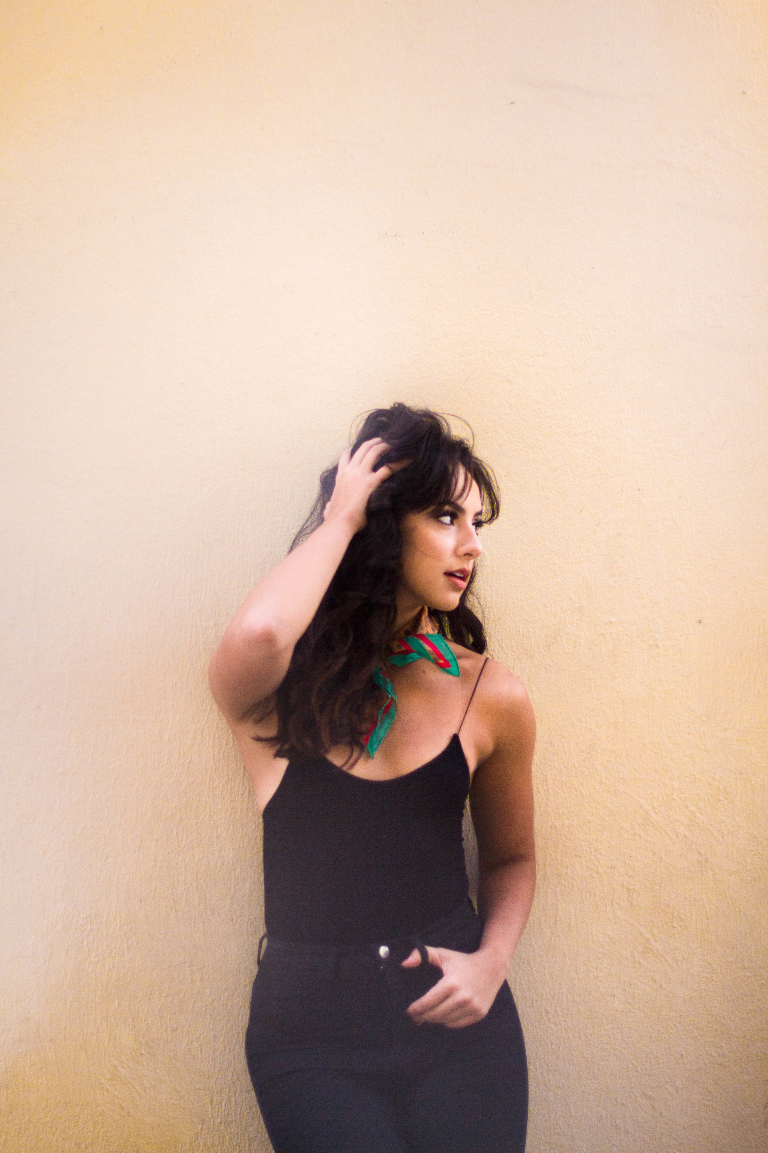  Woman standing infant of light yellow wall with her hand in her hair. She is wearing a colorful scarf around her neck and black tank top and jeans. 