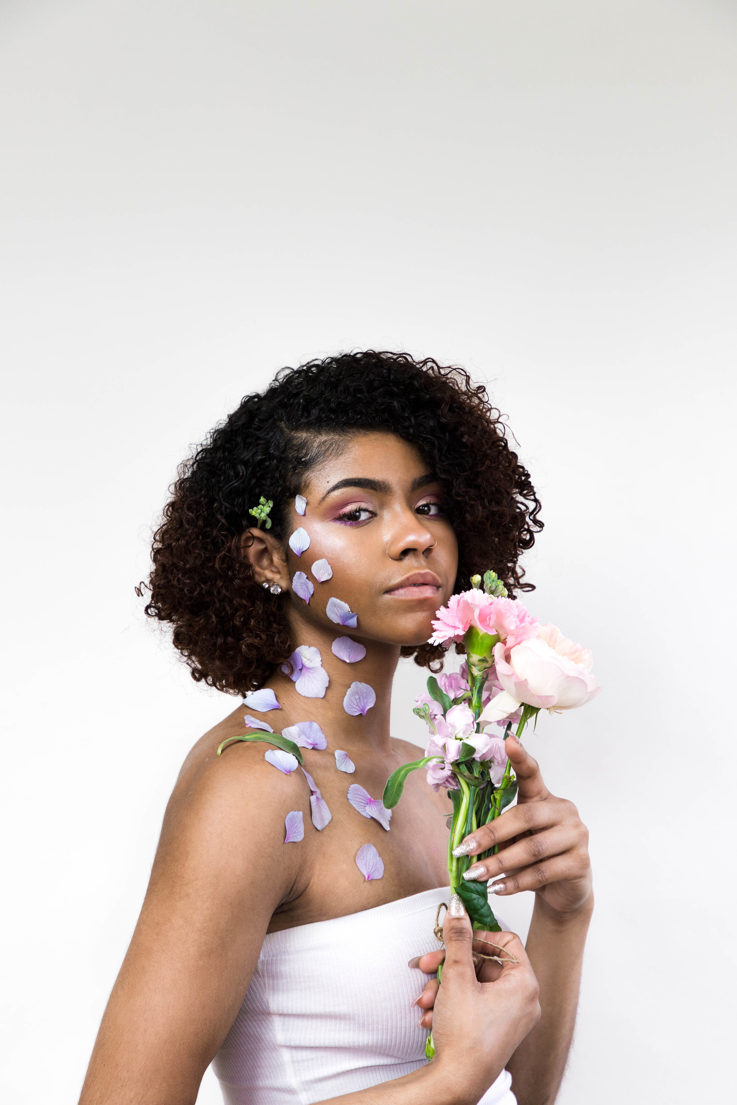  Woman with purple flower petals on her face and chest standing holding small bouquet of flowers against white backdrop. 