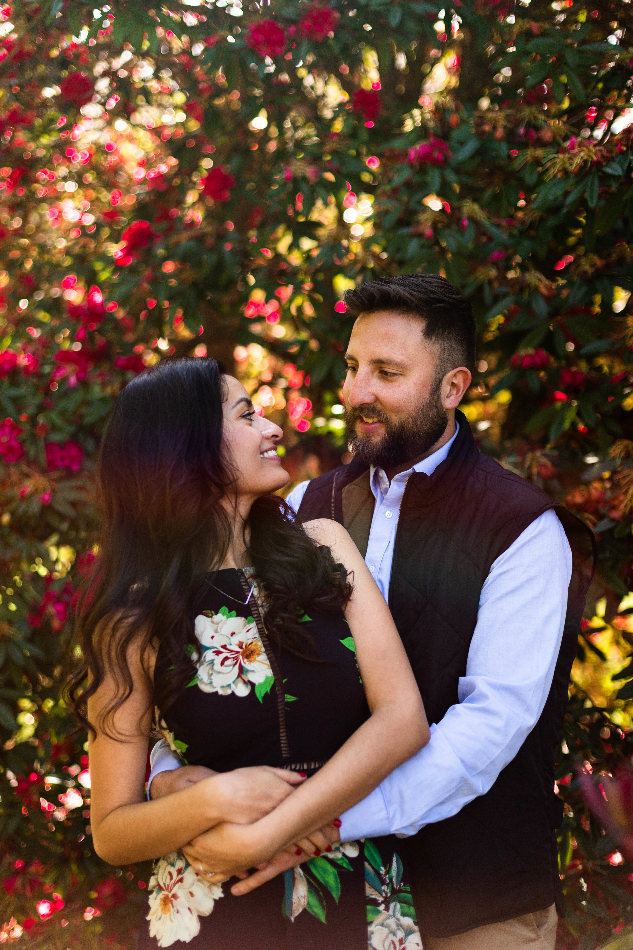  Couple in front of tree with red flowers. Woman with long black wavy hair wearing black dress with large white flowers. Man wearing light blue button down with a black vest. 
