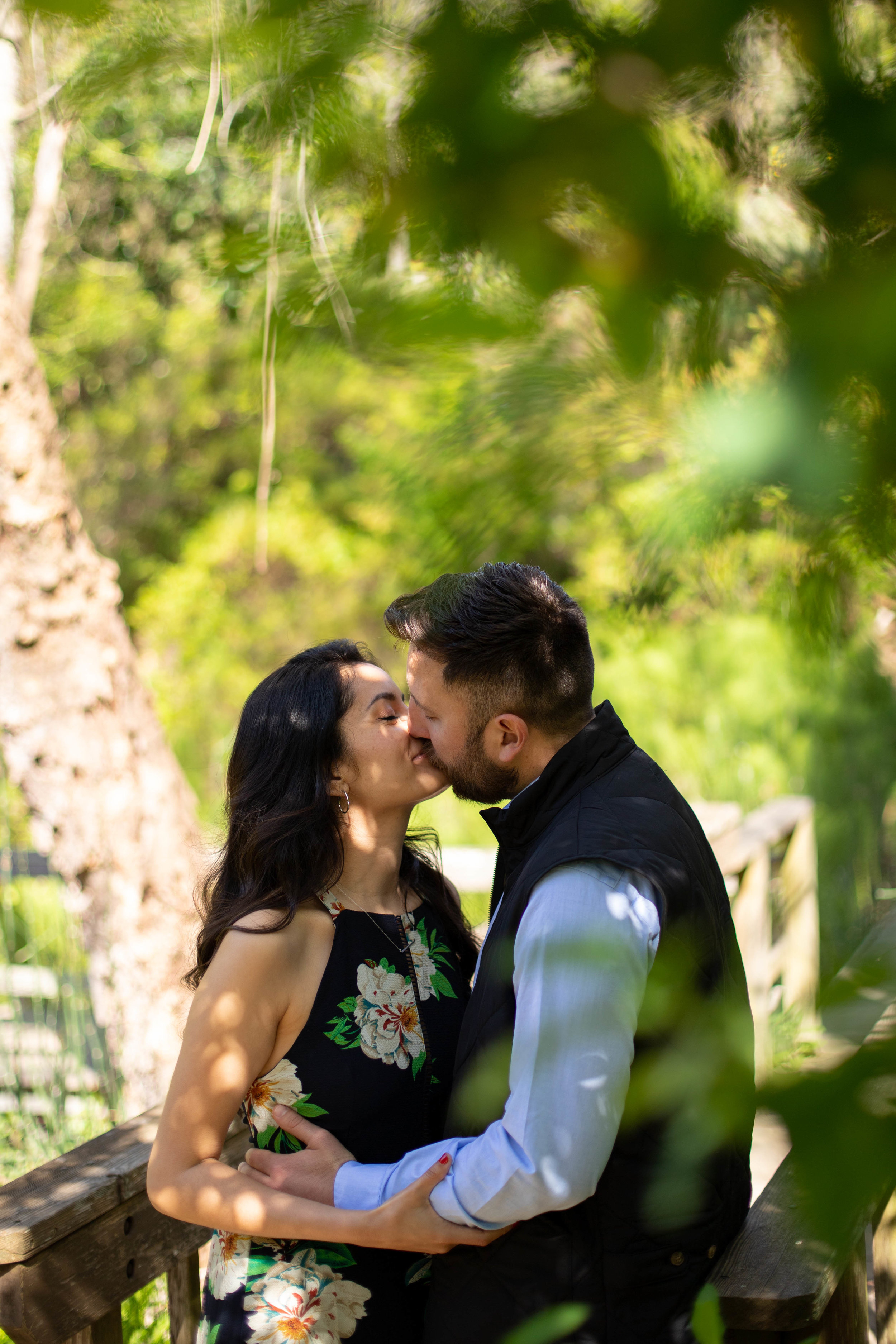  Couple kissing on a bridge. Leaves in the foreground. Woman is on the left; man is on the right with his hand on the small of her back. 