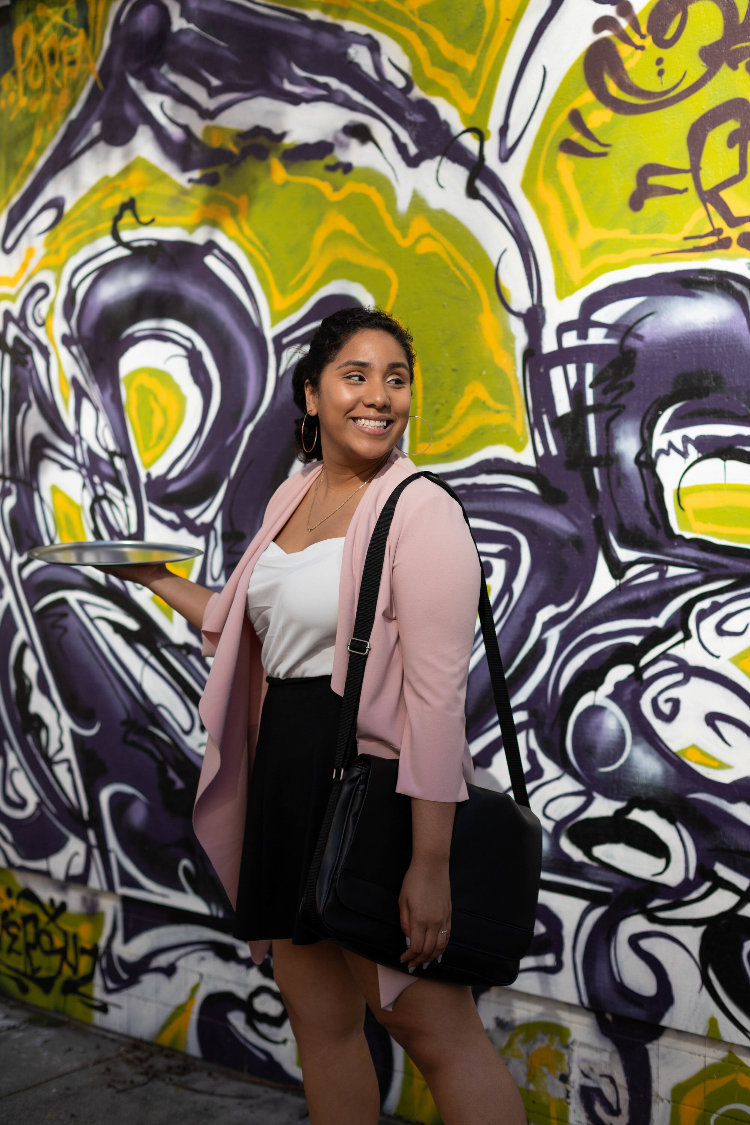  Woman in front of green and purple graffiti wall wearing pink cardigan, black skirt, and briefcase and holding serving tray walking and looking back over her shoulder. 