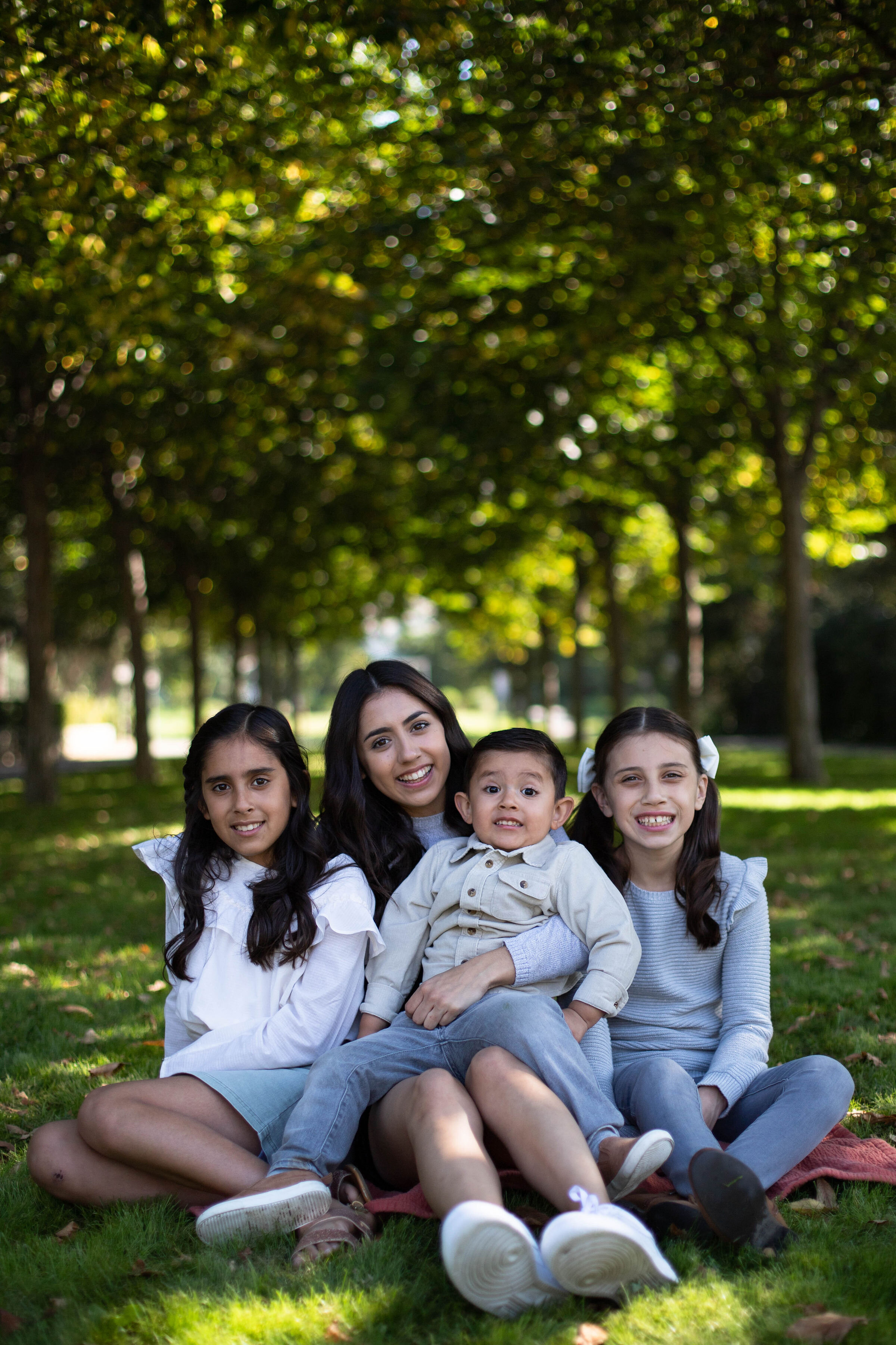  Four children wearing blue and white sitting on green grass in front of trees. Big sister holding littlest brother on her lap with two middle sisters on either side. All smiling and looking at camera. 