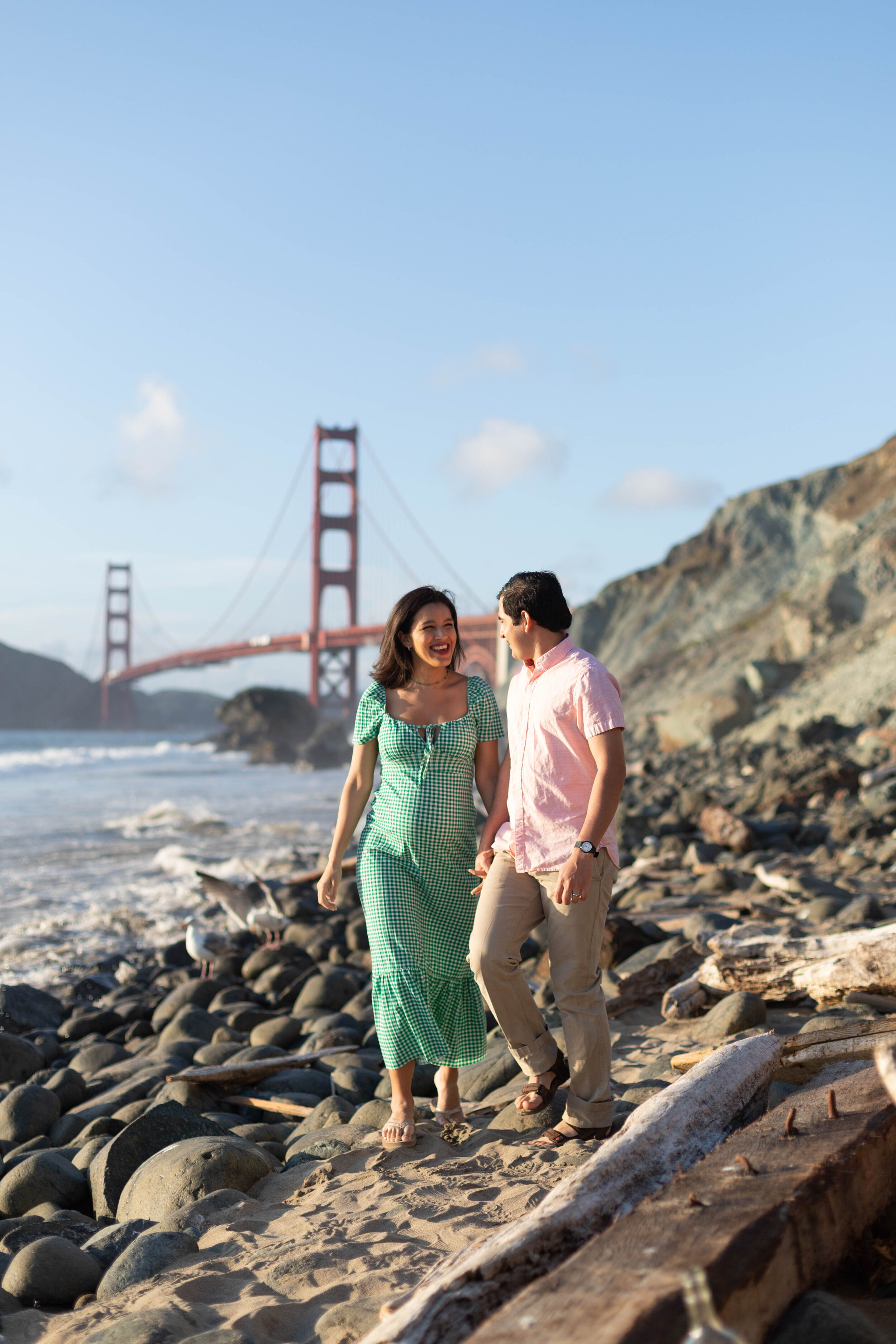  Pregnant couple walking on beach with Golden Gate Bridge in the background. Woman has short brown hair and is wearing a long, green, flowy dress. Man is wearing a pink shirt and khaki pants. They are looking at each other. 