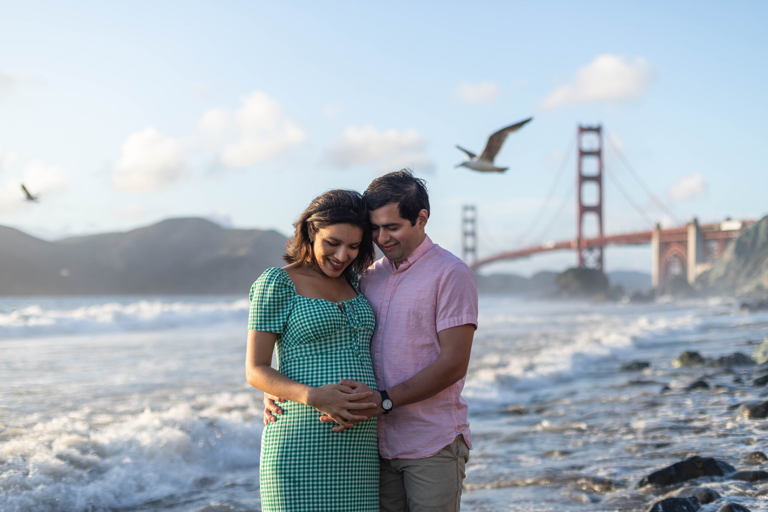  Pregnant couple standing together in front of Golden Gate Bridge with seagulls flying in the background. Couple looking down at woman’s pregnant belly while caressing it with their hands. 