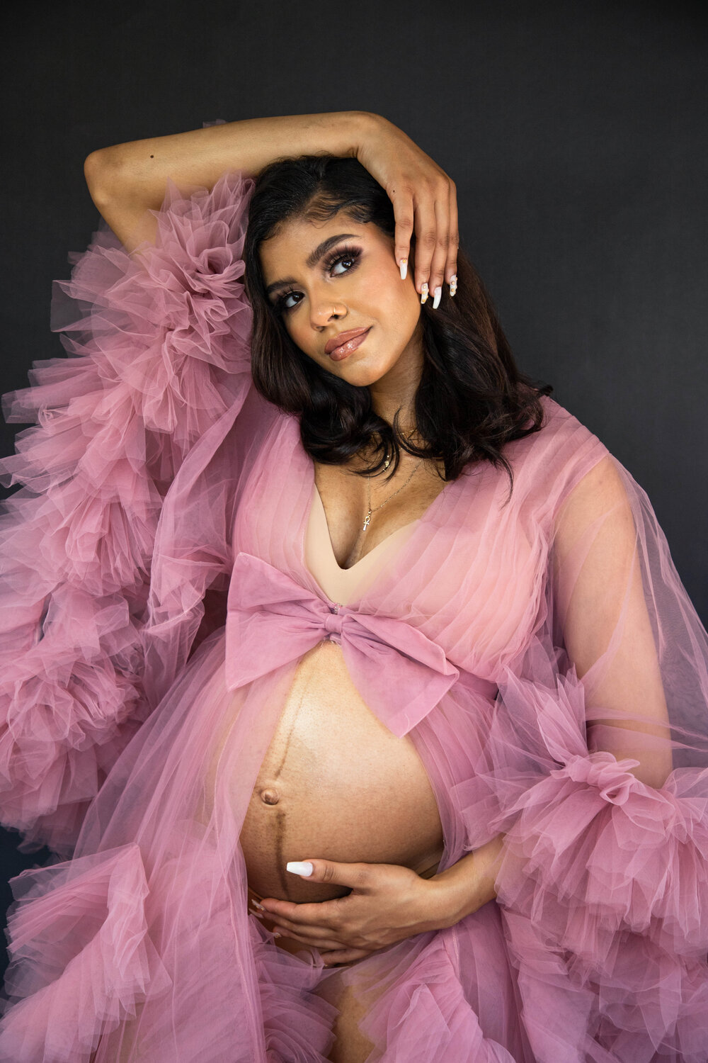  Pregnant woman wearing dusty rose ruffled robe with bell sleeves posing with one hand under belly and other hand on her head 