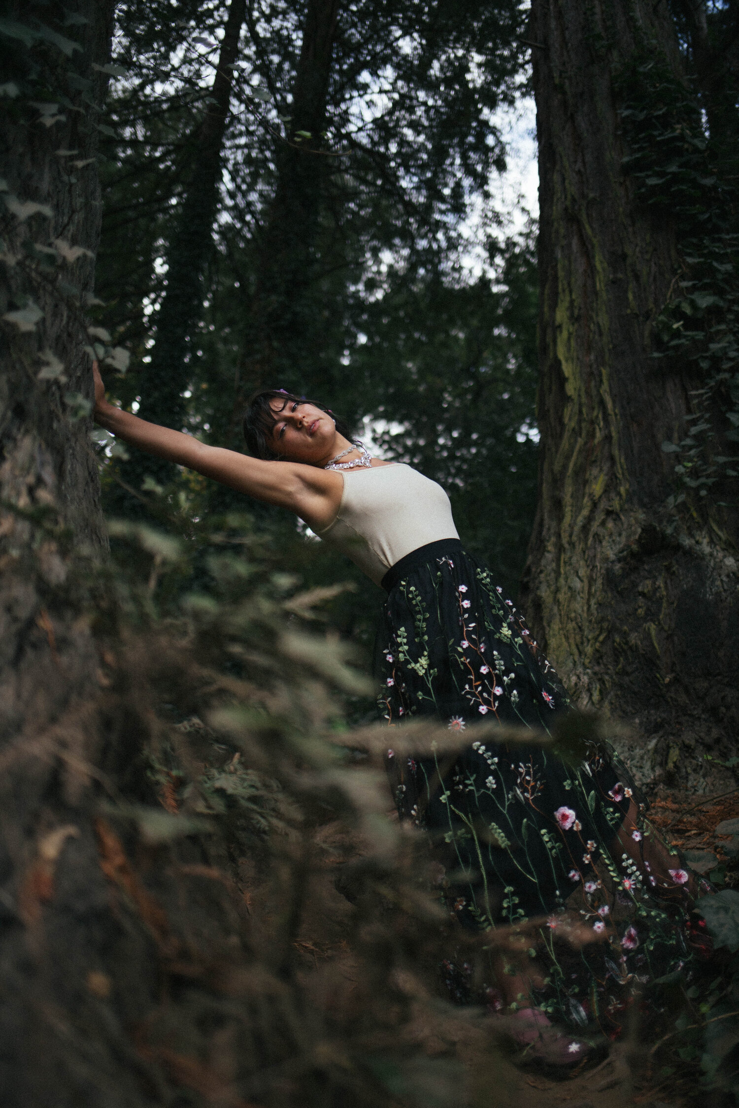  Woman leaning with arm against redwood tree wearing light tank top and long black embroidered skirt. Branches in the foreground 