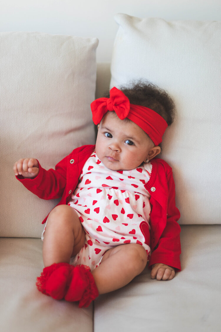  Baby girl wearing red cardigan, socks, and headband and white dress with pink and red hearts. She is sitting on a white couch and she has one hand up. 