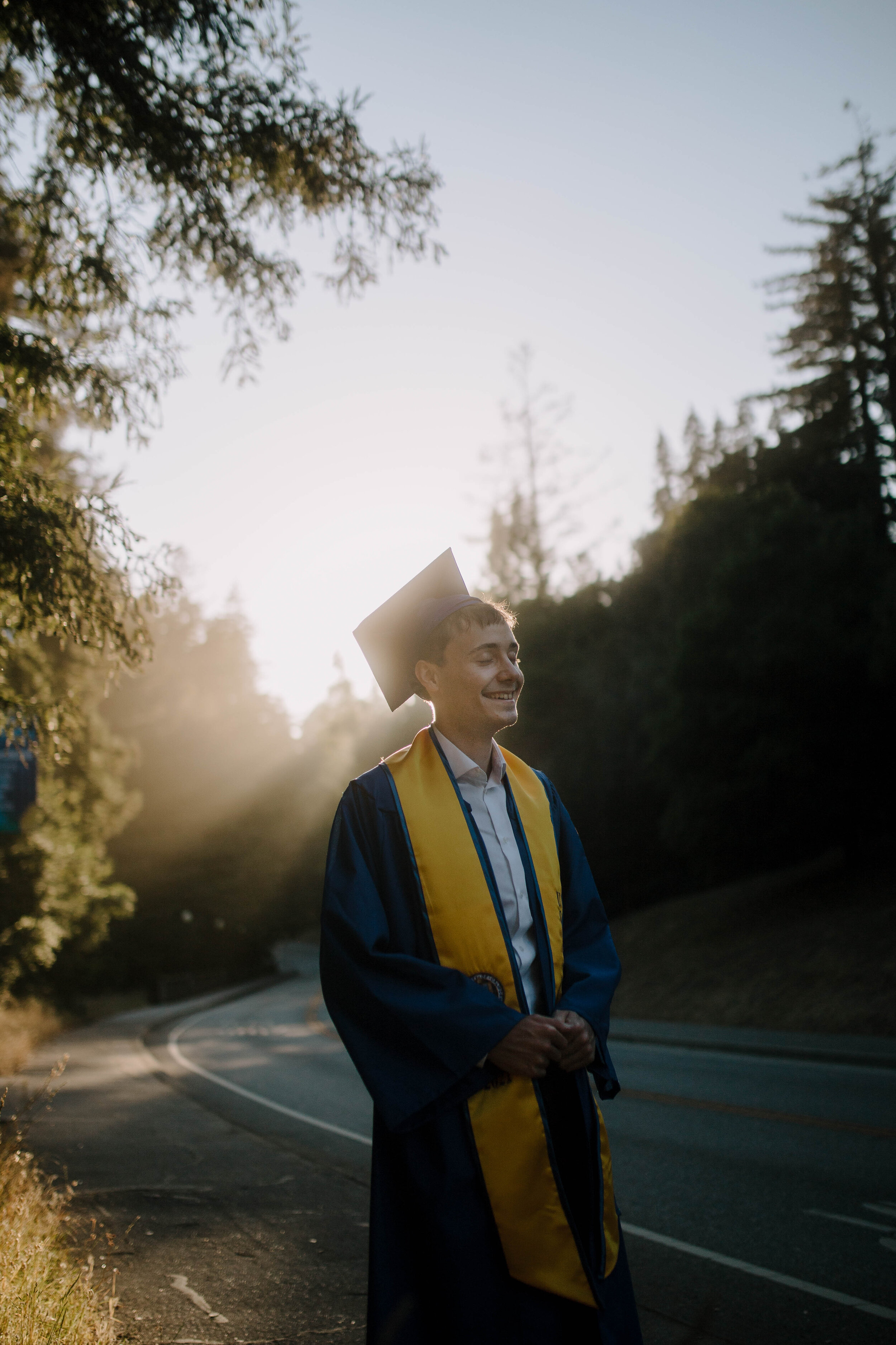  Graduate wearing blue robe and yellow stole in front of road with redwoods on either side. Light streaming behind the person to create a halo effect. 