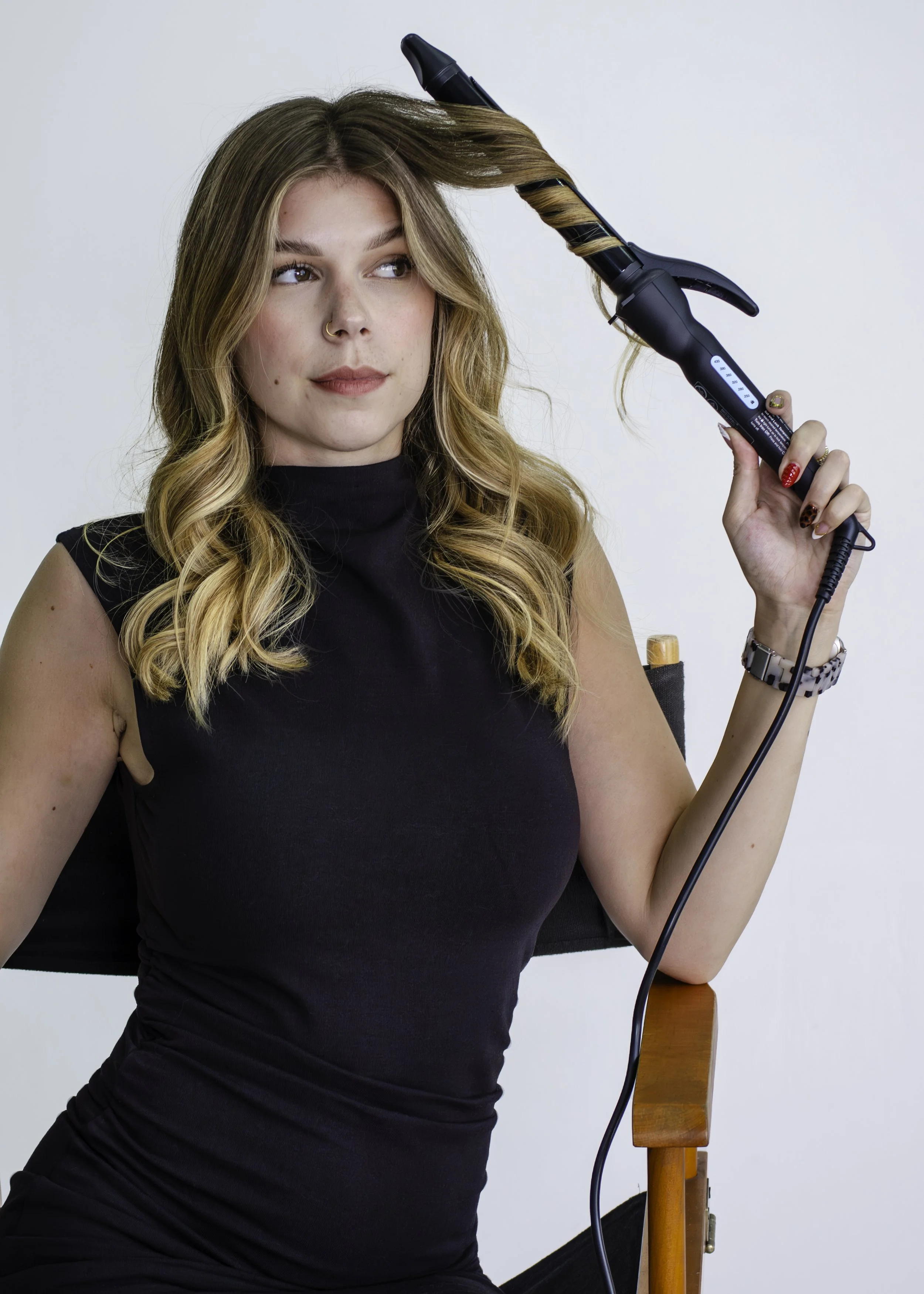 A woman with wavy blonde hair and a nose piercing is sitting on a wooden chair, using a curling iron to curl her hair. She is wearing a sleeveless black top and has red and leopard-print nails.