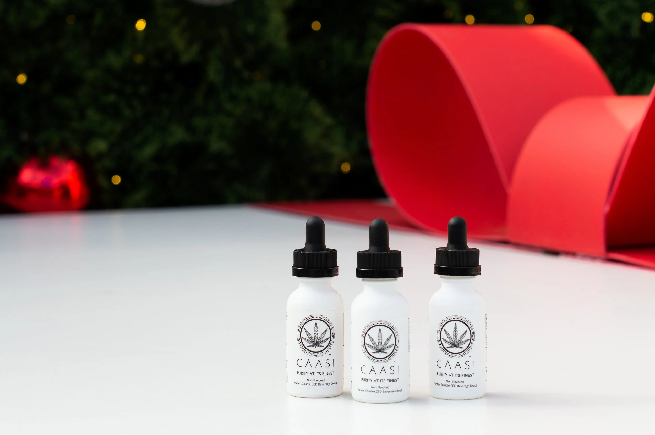 Three bottles of Caasi CBD drops on a white surface, with a blurred green Christmas tree and red decorations in the background.