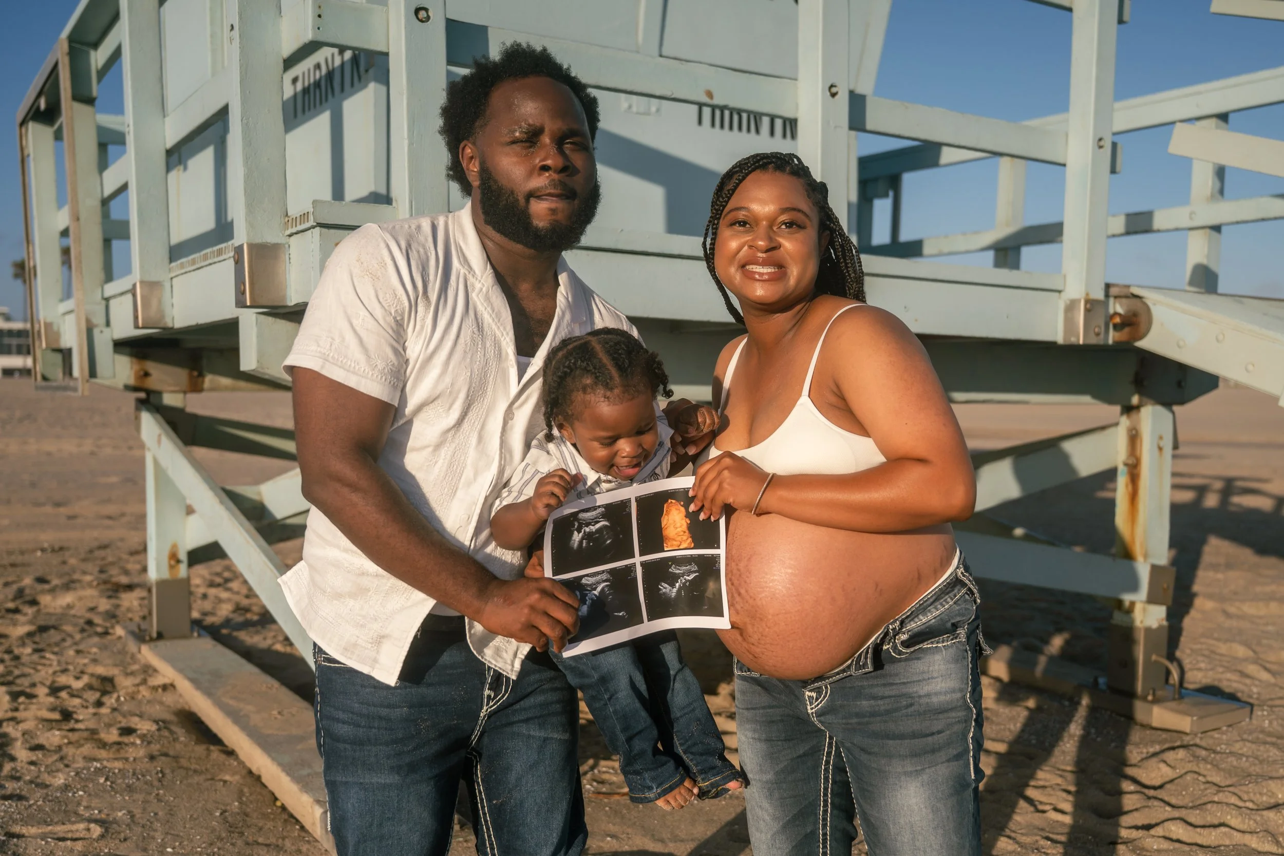 A happy family of three on the beach, with a pregnant woman, a man, and a young child, holding ultrasound images. They are standing in front of a beach lifeguard tower under a clear blue sky.