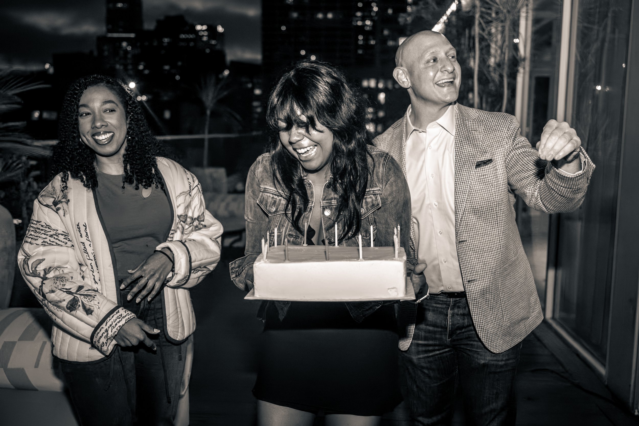 A group of three people celebrating a birthday at night, with a cake with candles, standing outside in an urban area with buildings in the background. The group includes a woman with curly hair, a woman with straight hair, and a man wearing a checkered blazer. The man is smiling and pointing.