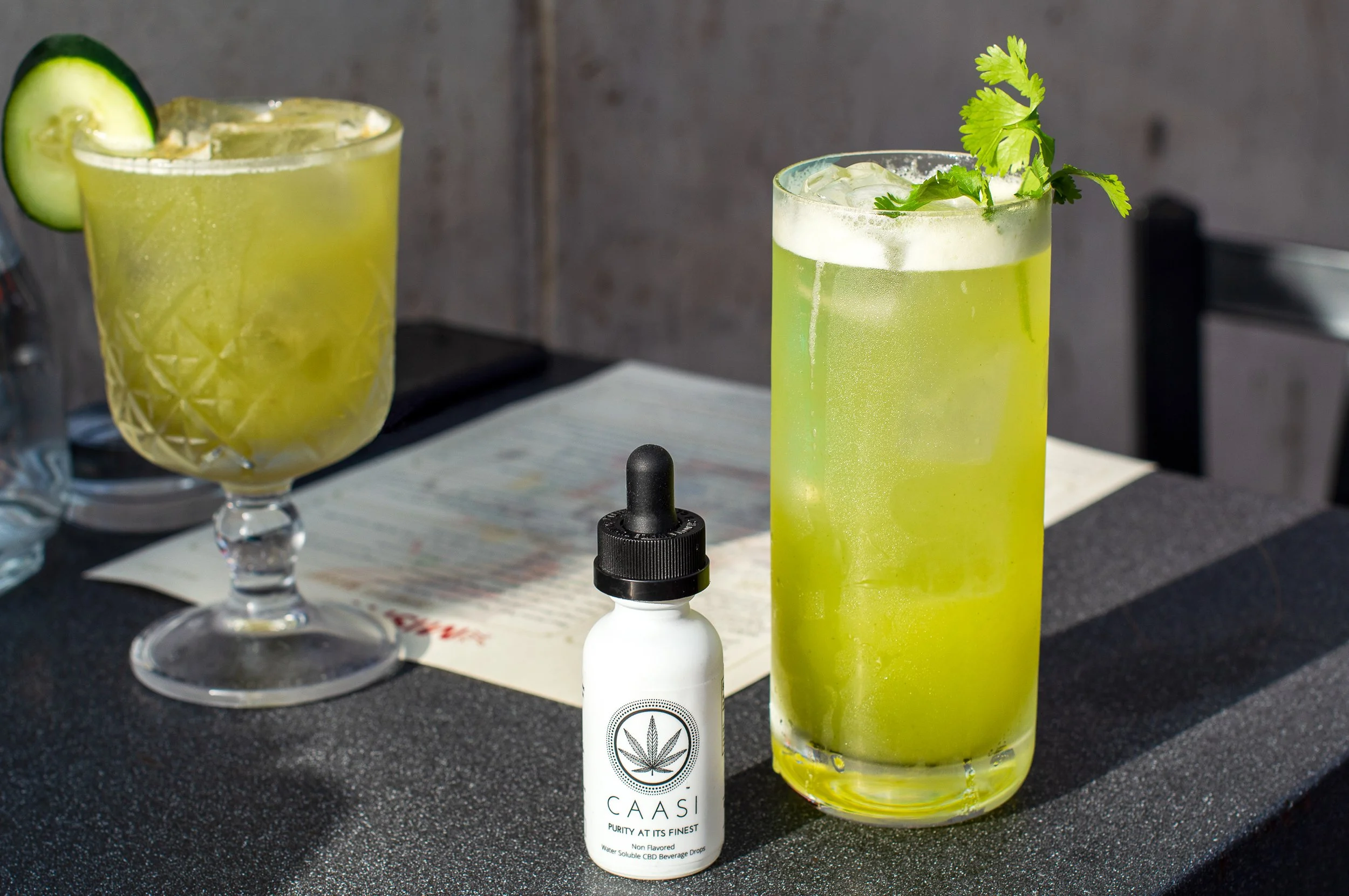 Two green cocktails with ice and garnish, one with a cucumber slice, on a dark table with a menu and a CBD tincture bottle nearby.
