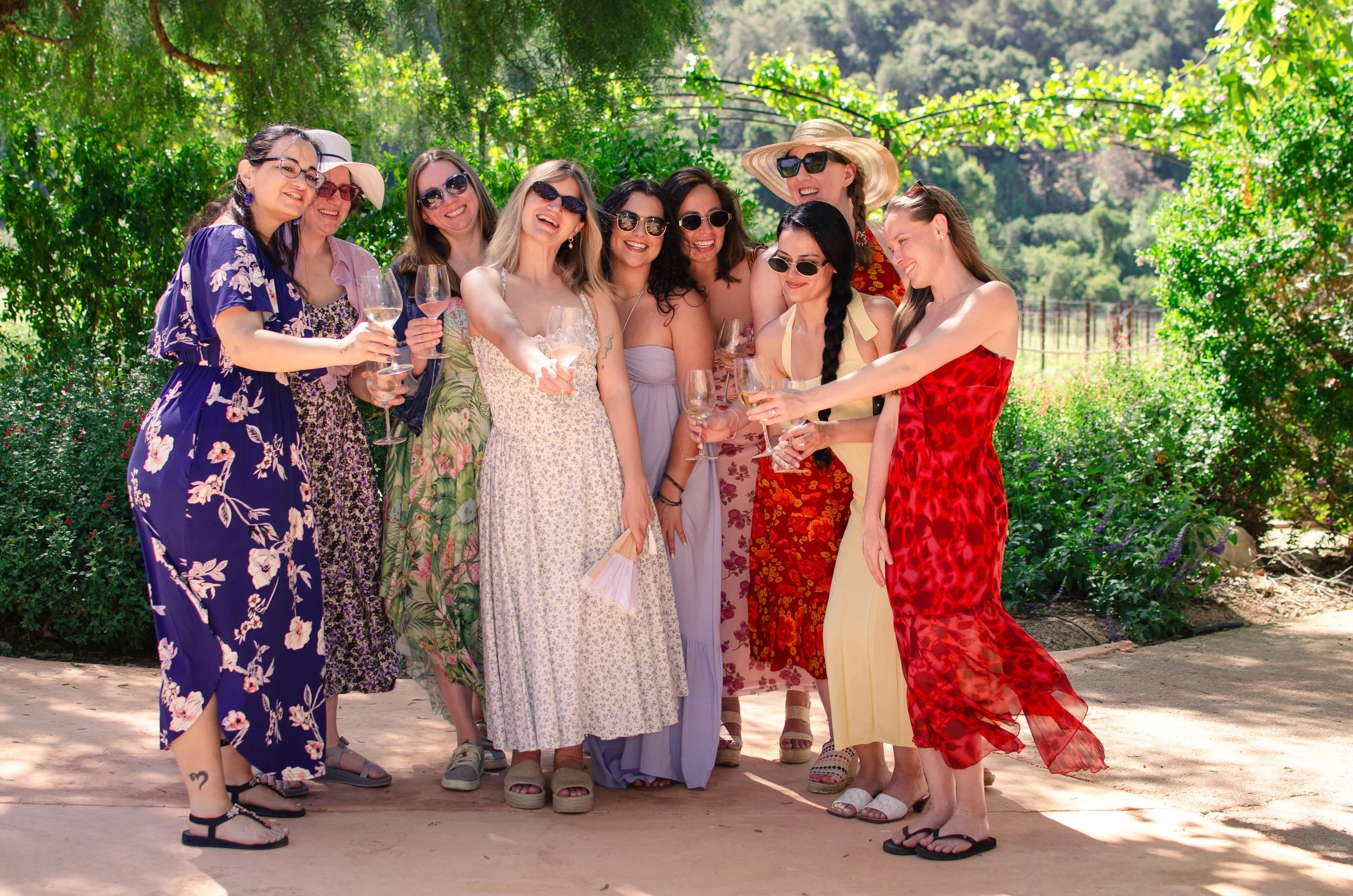 A group of women in colorful dresses and sunglasses enjoying a sunny outdoor gathering, holding wine glasses and smiling.