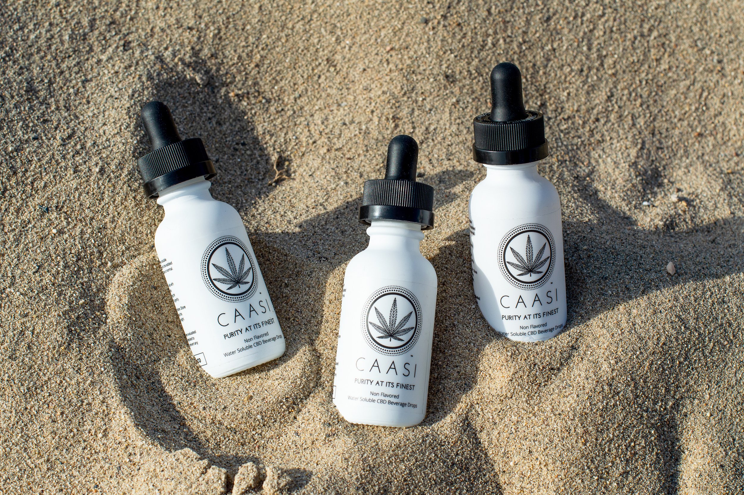 Three white bottles with black dropper caps lying on sandy beach, each labeled CAASI with a cannabis leaf logo, advertising CBD beverage drops.