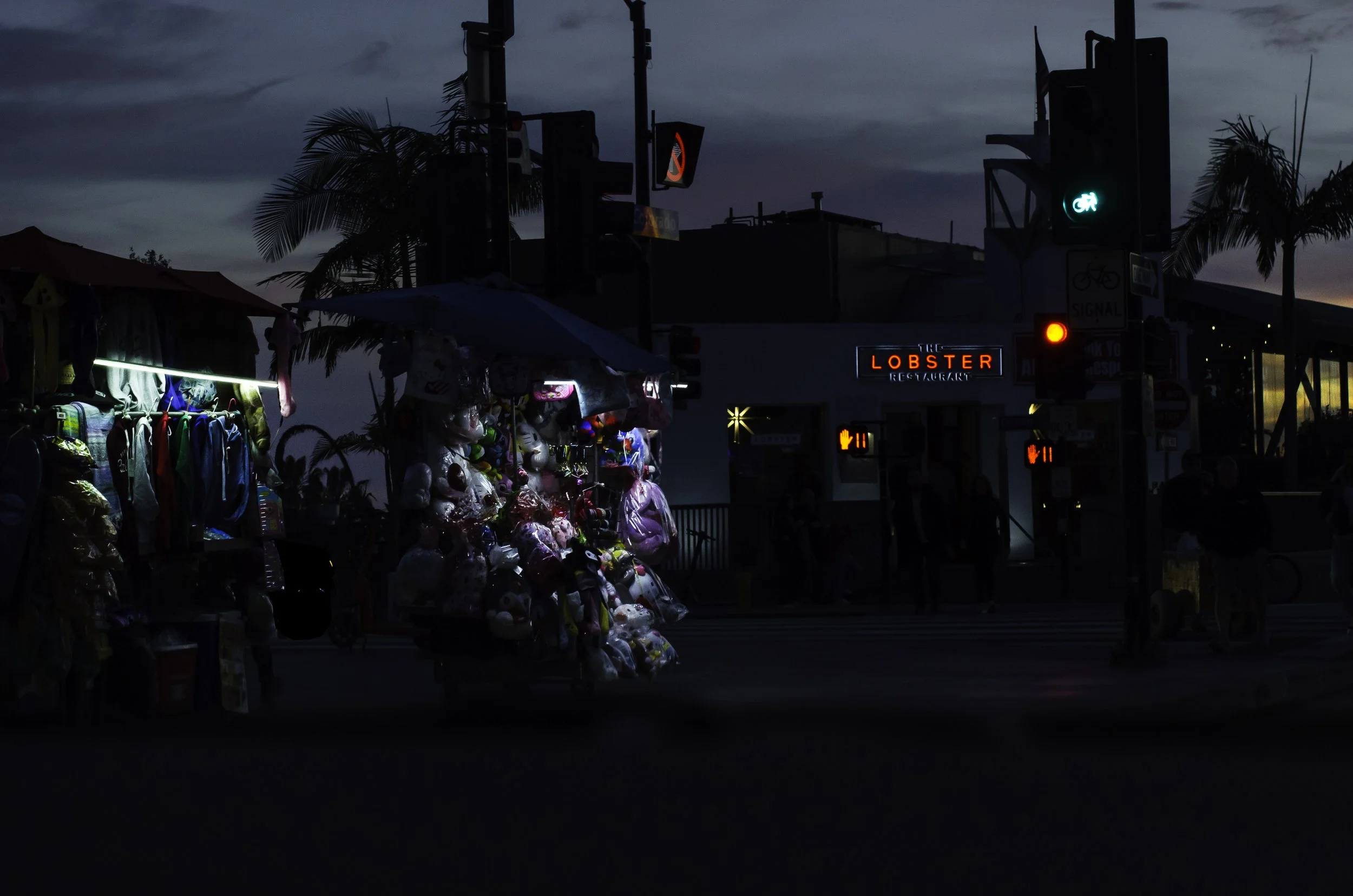 Street vendor selling plush toys and clothing at dusk, with a restaurant called 'The Lobster' in the background, palm trees, and traffic signals.