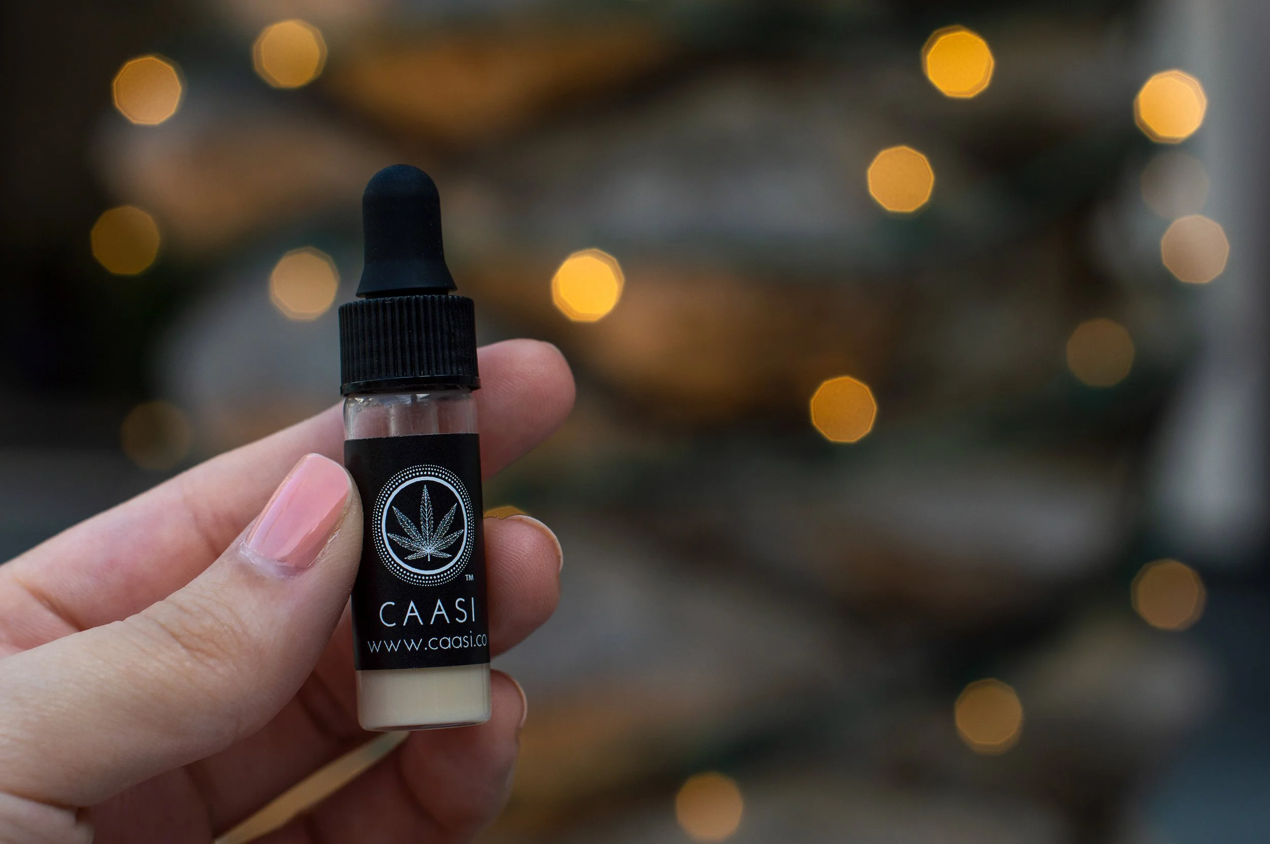 Hand holding a small bottle labeled 'CAASI' with a cannabis leaf logo, with blurred warm lights in the background.