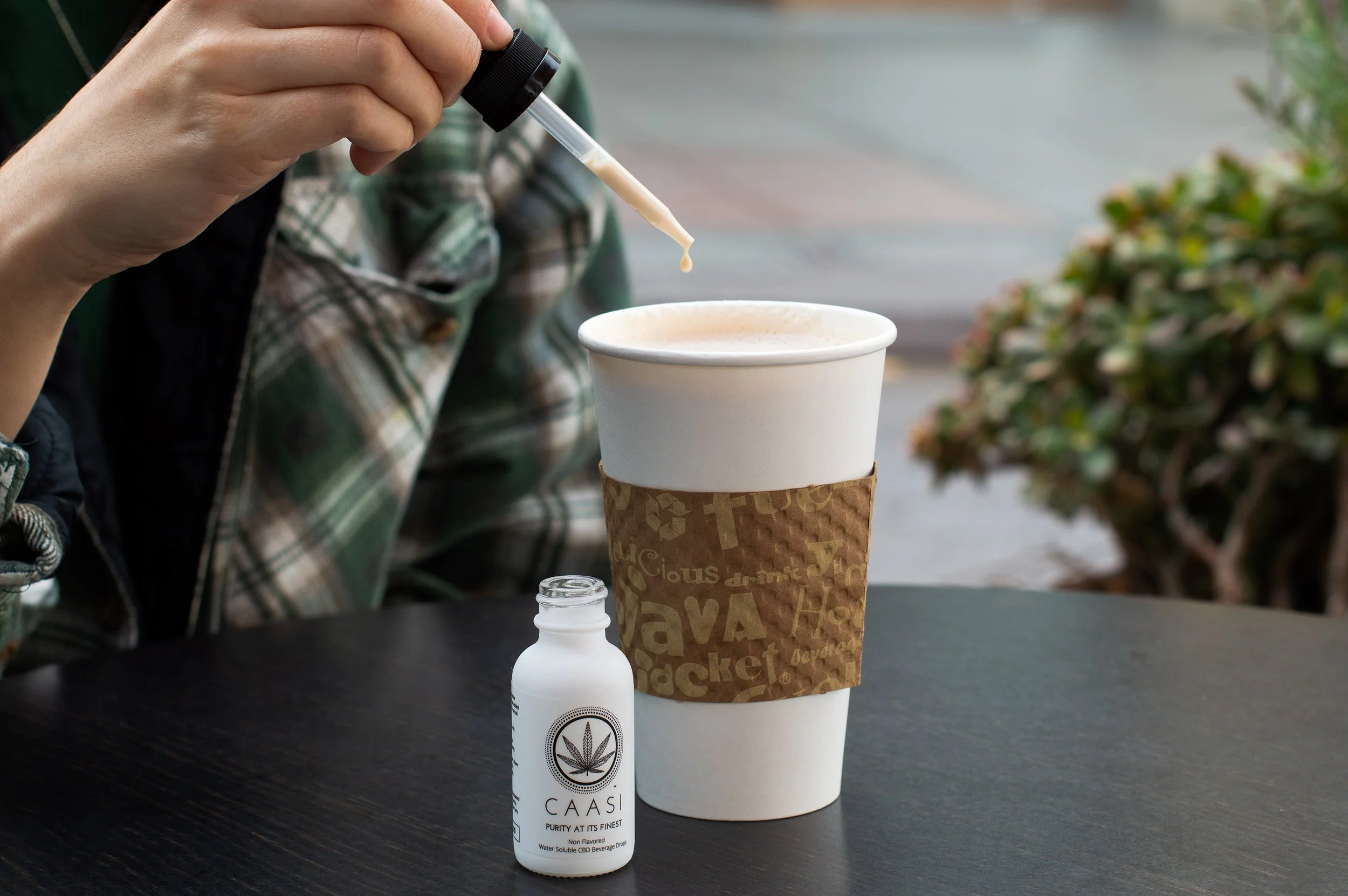 A person holding a dropper above a to-go coffee cup on a table, with a CBD product bottle nearby.