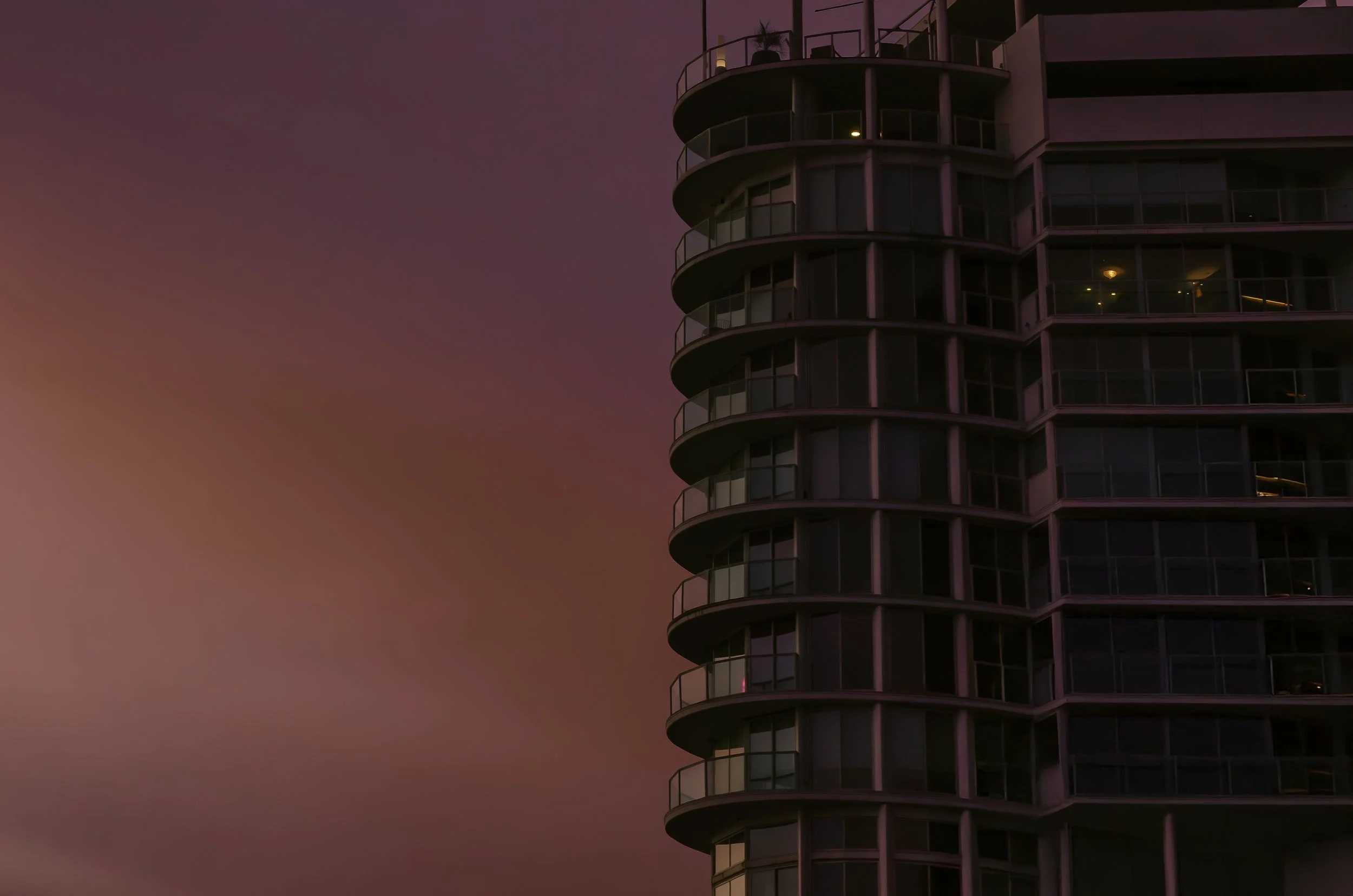 Close-up of a modern high-rise building with curved balconies and glass windows, against a pinkish-purple sunset sky.