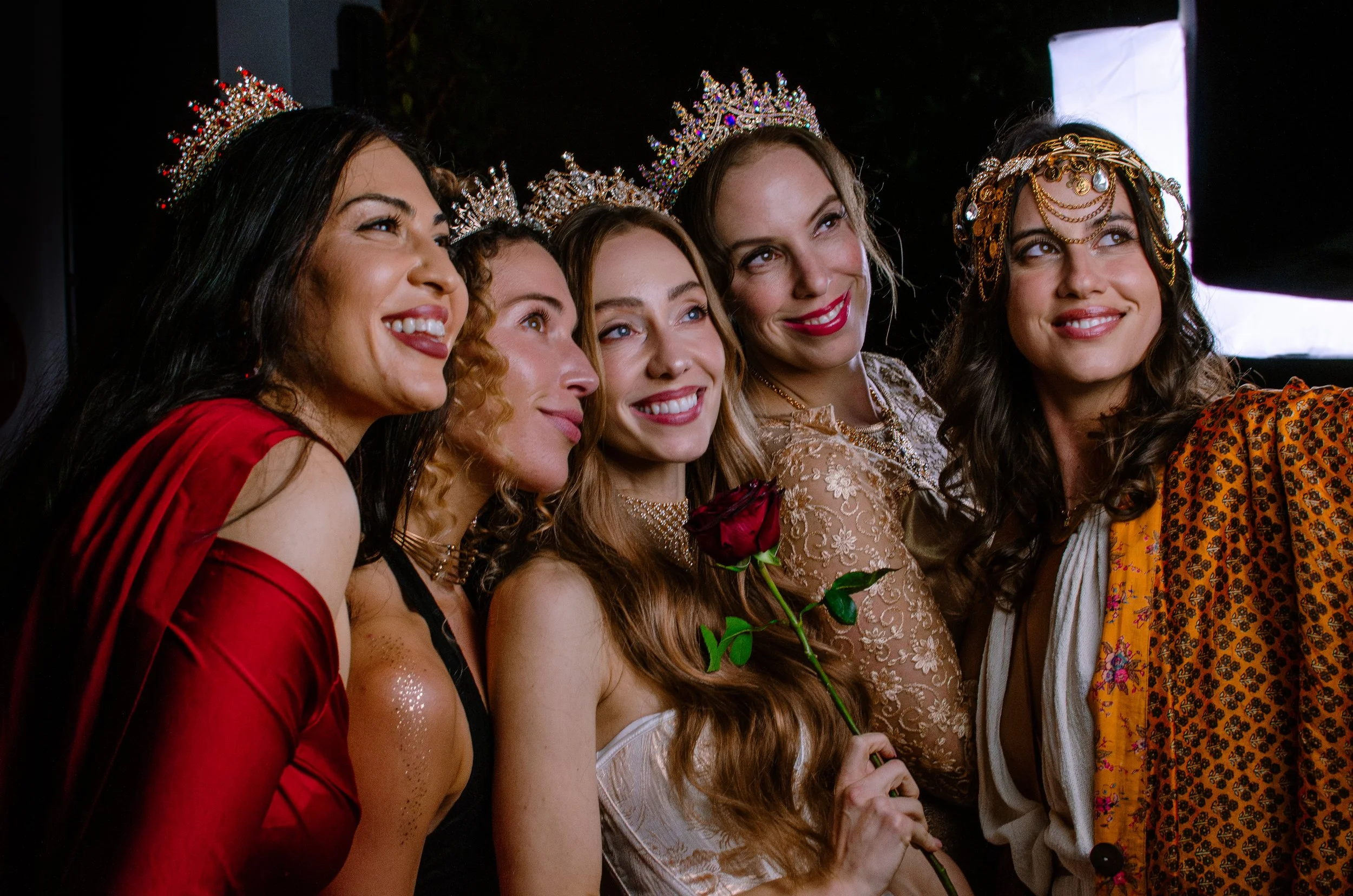 Group of five women wearing tiaras, smiling, dressed festively, some holding flowers, taking a selfie or posing together in an event.