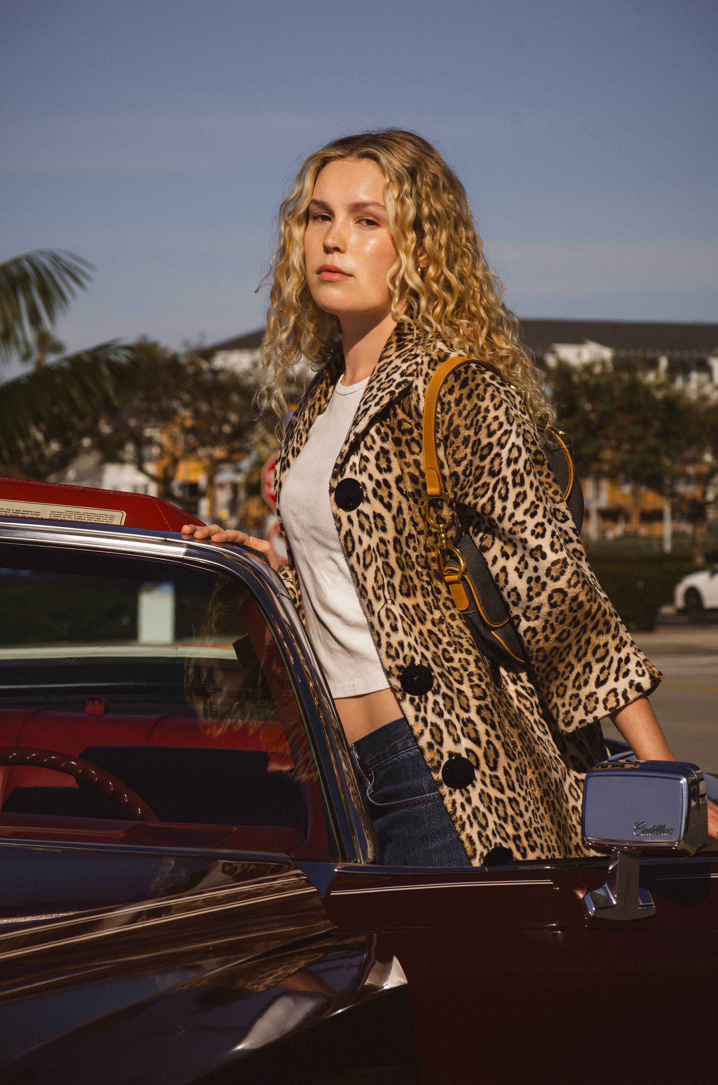 A woman with curly blonde hair wearing a leopard print coat, white t-shirt, and jeans, standing next to a red vintage Cadillac with palm trees in the background.