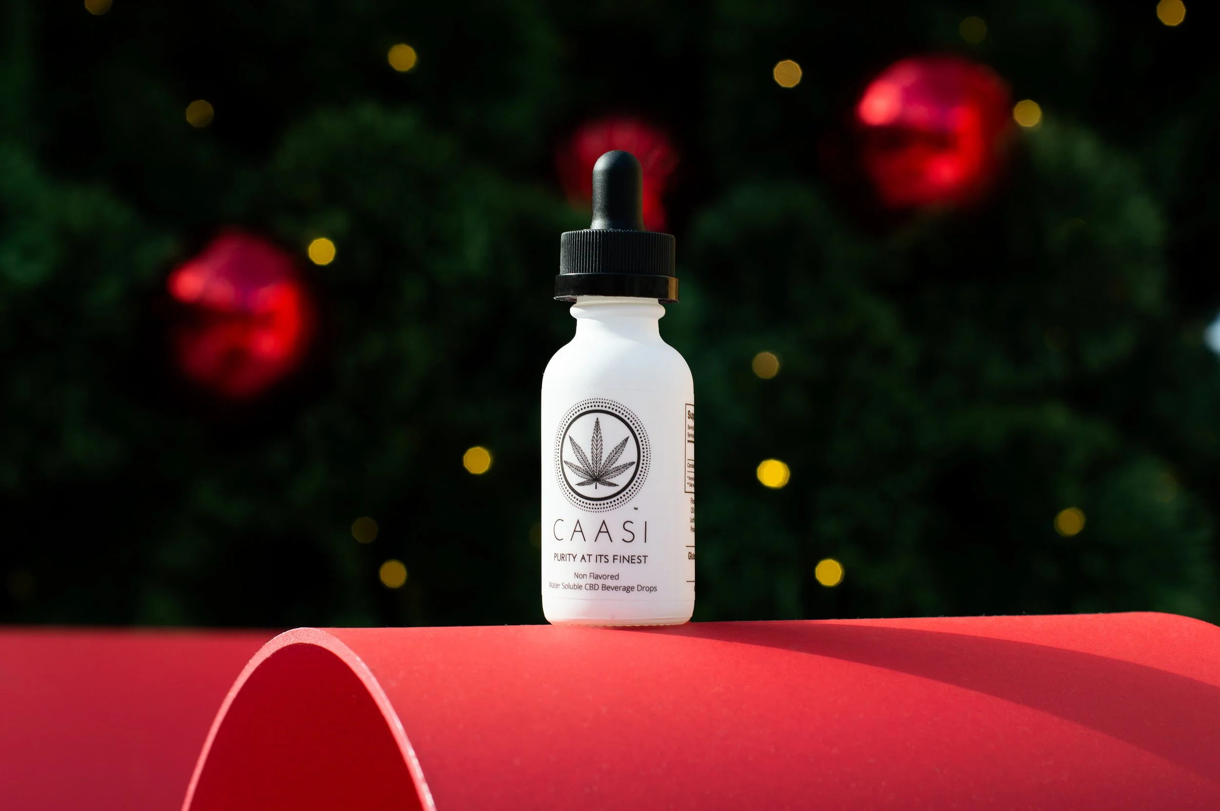 A white bottle with a black dropper cap labeled CAASI, featuring a leaf symbol, set on a curved red surface in front of a blurred Christmas tree with red ornaments and yellow fairy lights.