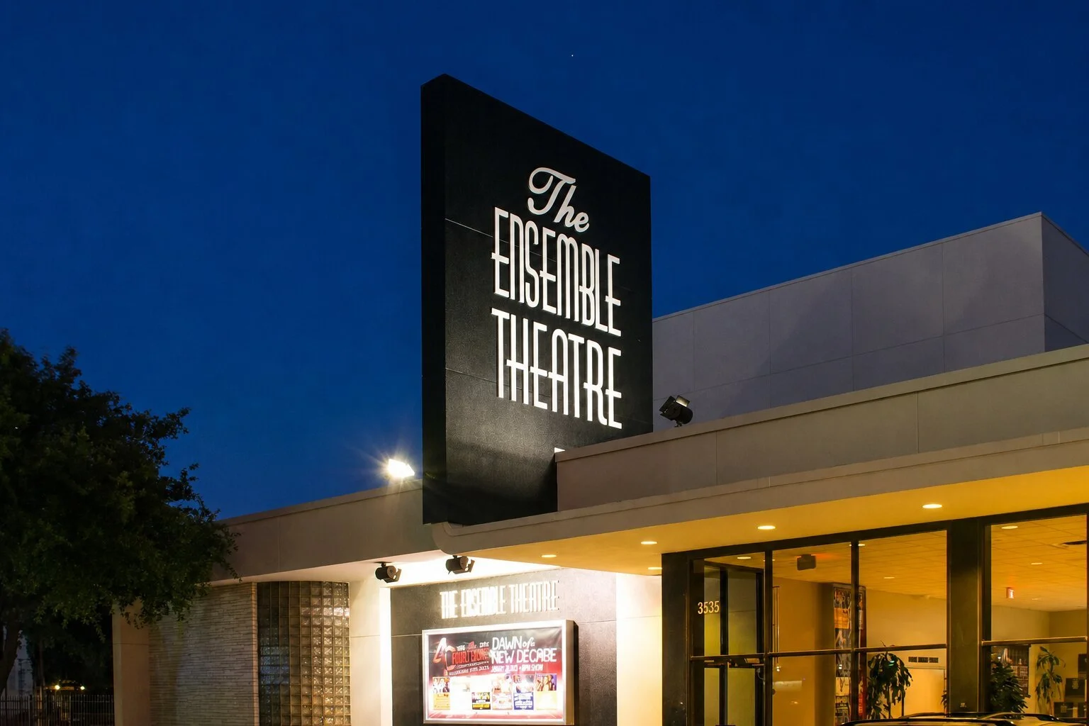 The Ensemble Theatre in Houston at night with a tall illuminated exterior sign above the entrance and warmly lit building facade.